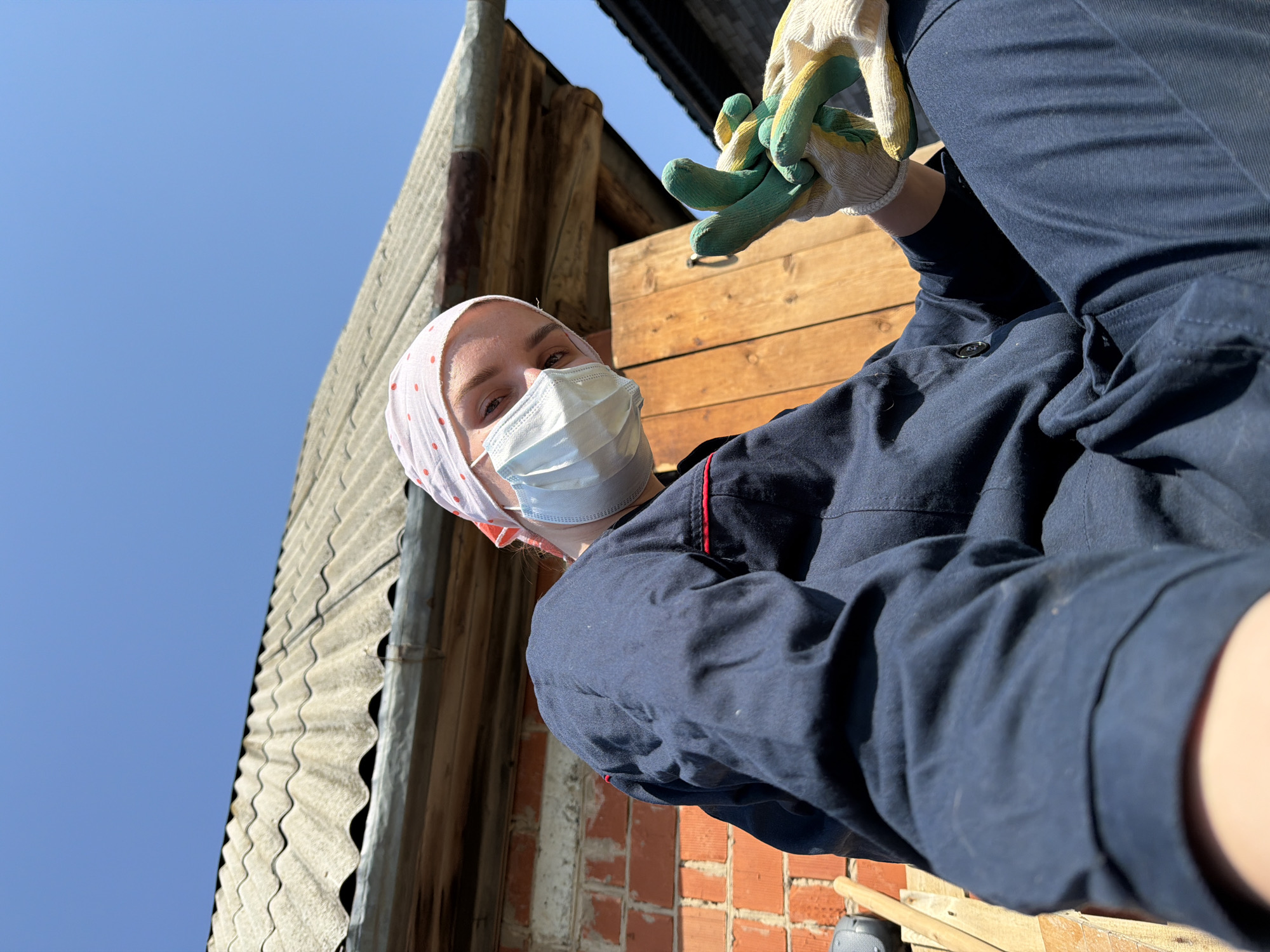 A person wearing a headscarf, a light blue surgical mask, and a dark blue jacket is holding up work gloves. The person is standing outside next to a building with corrugated metal siding and a brick wall. The sky is bright blue. Based on the information provided, the location is Chaikovsky, Russia.
