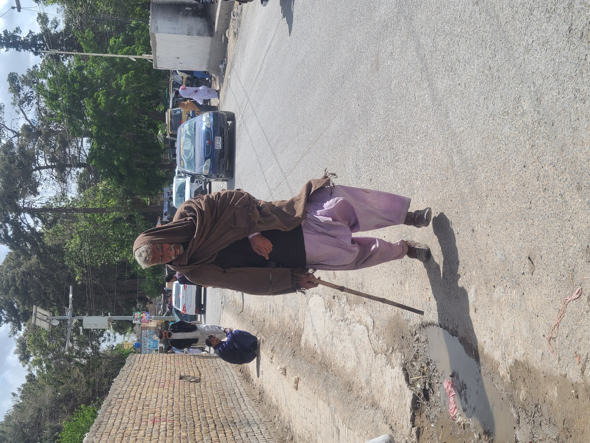 A middle-aged man in Quetta City Tehsil, Pakistan, walks down a street with a brown shawl, lavender pants, and a wooden walking stick. A blue car is parked on the street. There is a brick wall along the sidewalk and a puddle on the ground next to the man. Other people are in the background.
