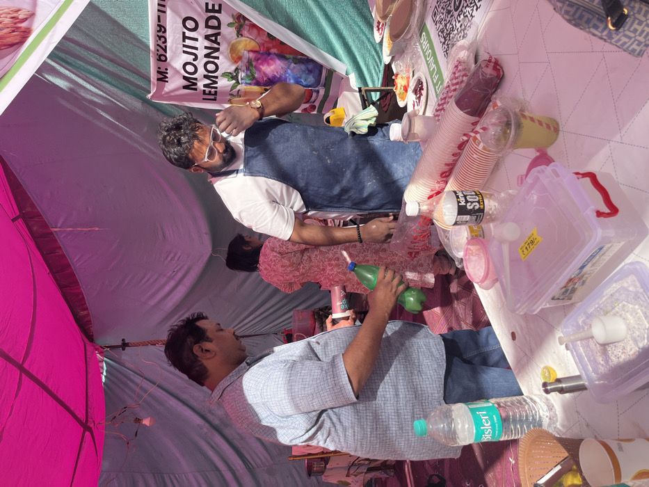 The scene is a food stall in a market in Chandigarh, India, at the coordinates 30.764, 76.798. There are three people in the scene. One man, wearing a plaid shirt, is holding a green bottle and pouring liquid into a container. Another man, wearing a white shirt and a blue apron, is wiping his face with a napkin. A woman in a pink shirt is standing in the background. The stall has a pink and white canopy, a table with various food items, containers, and beverages. The table has a sign that reads "Mojito Lemonade," and there are signs with a QR code and the words "Fruit Flavours." There are empty cups, a bottle of water, and a bottle of soda on the table. The scene appears to be a busy street market in Chandigarh, India.