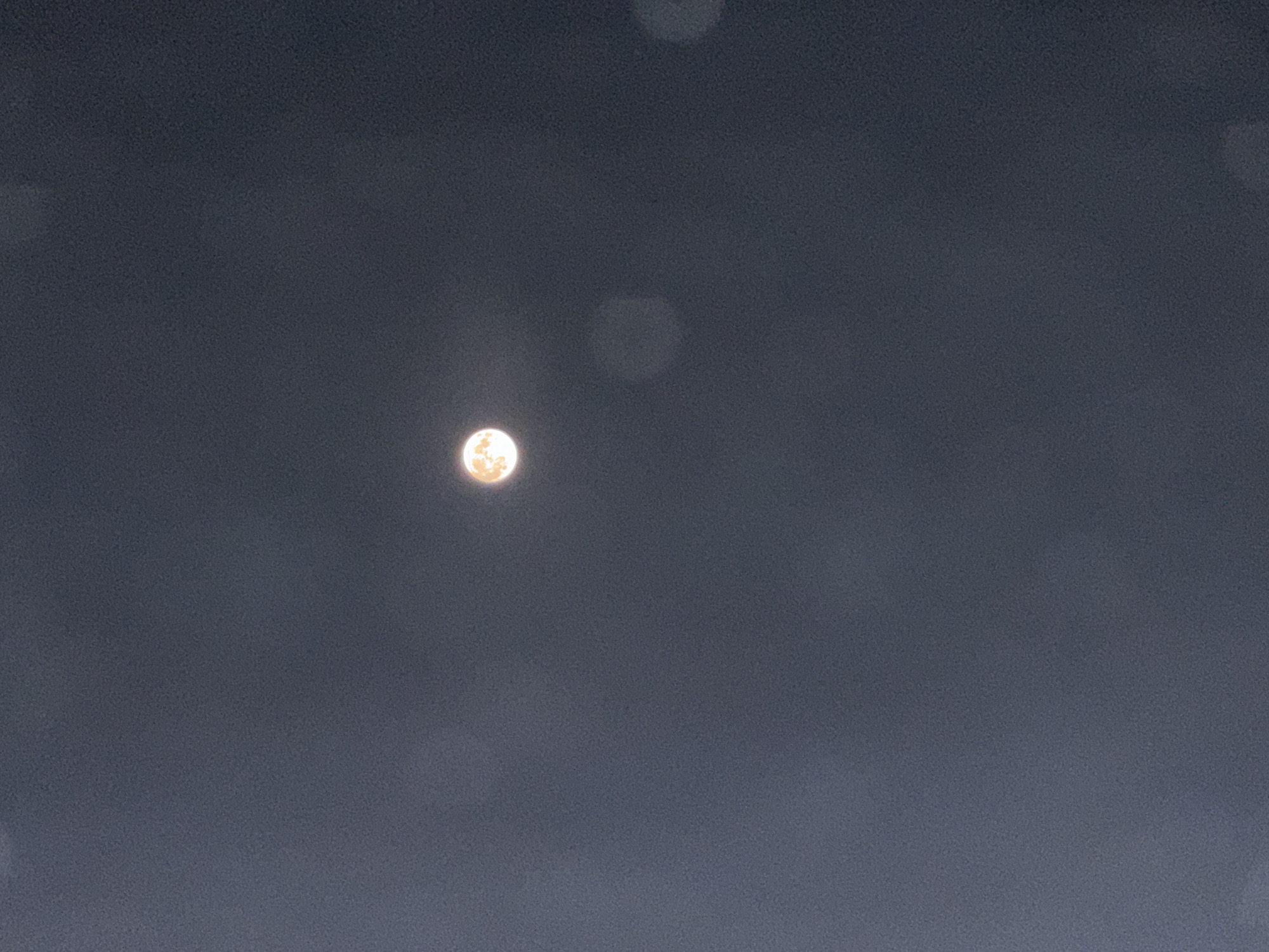 The image shows a full moon in a dark, hazy sky. The moon is a bright, orange-yellow orb, and it appears to be surrounded by a faint halo. There are several blurry, circular lights in the foreground, which could be reflections on a window or lens flare. The overall impression is one of a slightly obscured view of the moon. The location in Zatoka, Ukraine, is not discernible from the image itself.