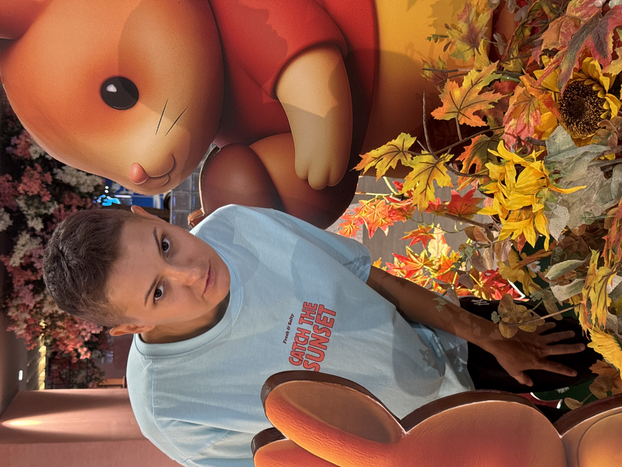 A young person with short, dark hair and a light blue t-shirt that says "Fresh & Salty CATCH THE SUNSET" in red lettering, leans towards the camera. They are positioned to the left of a large, cartoonish orange rabbit-like character. The rabbit is holding a brown, acorn-shaped object. The background consists of a display of artificial autumn foliage, featuring vibrant red, orange, and yellow leaves, as well as sunflowers. The lighting suggests an indoor setting, possibly a themed display in a shopping mall or entertainment venue. There is no indication of Nha Trang Ward, Vietnam in the image.