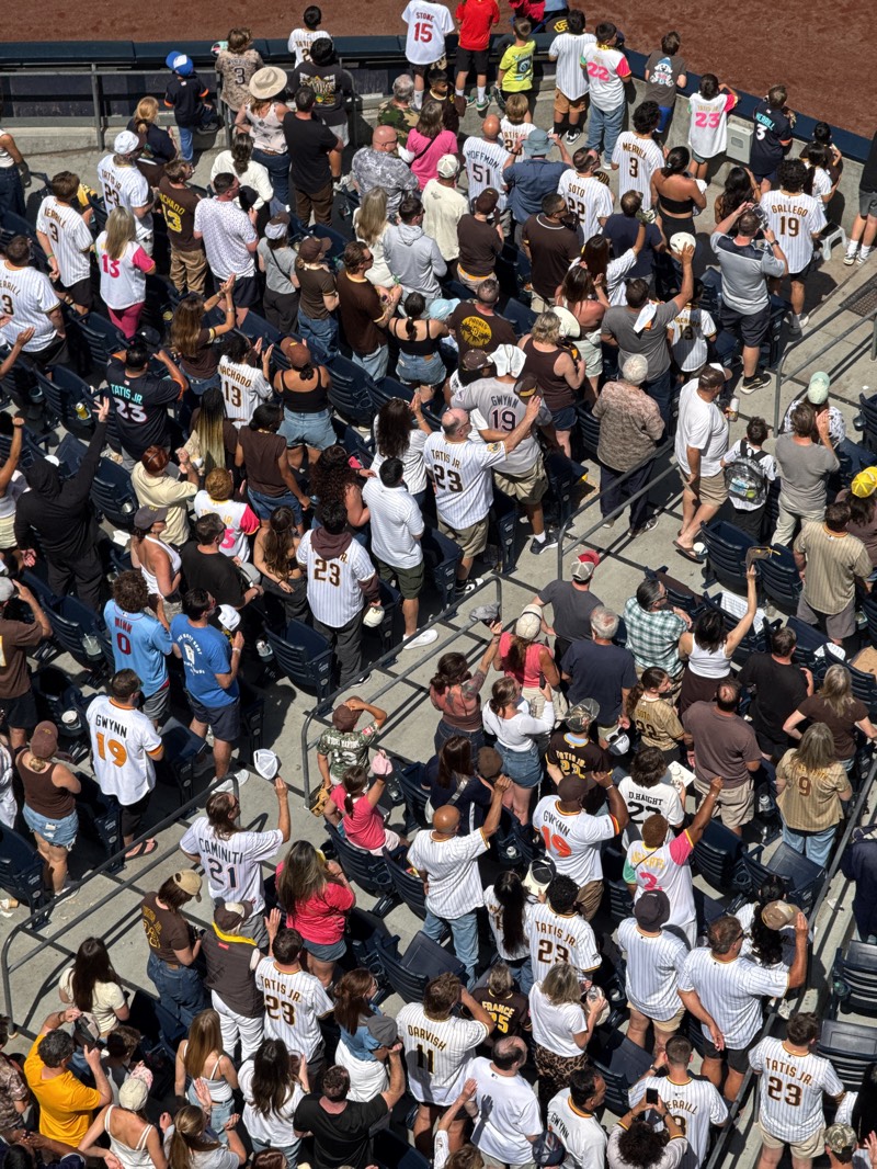 The image depicts a large crowd of baseball fans in the stands, likely at Petco Park in San Diego, United States. The fans are densely packed in seats, with many wearing Padres jerseys, particularly those of players like Tatis Jr., Gwynn, and Machado. Some are standing, raising their arms, cheering, or holding hats. The scene is brightly lit, indicating it is during the day, and the view is from an elevated position, providing an overview of a section of the stadium. The environment consists of typical stadium seating and a mix of people of various ages and appearances, mostly facing towards the baseball field.