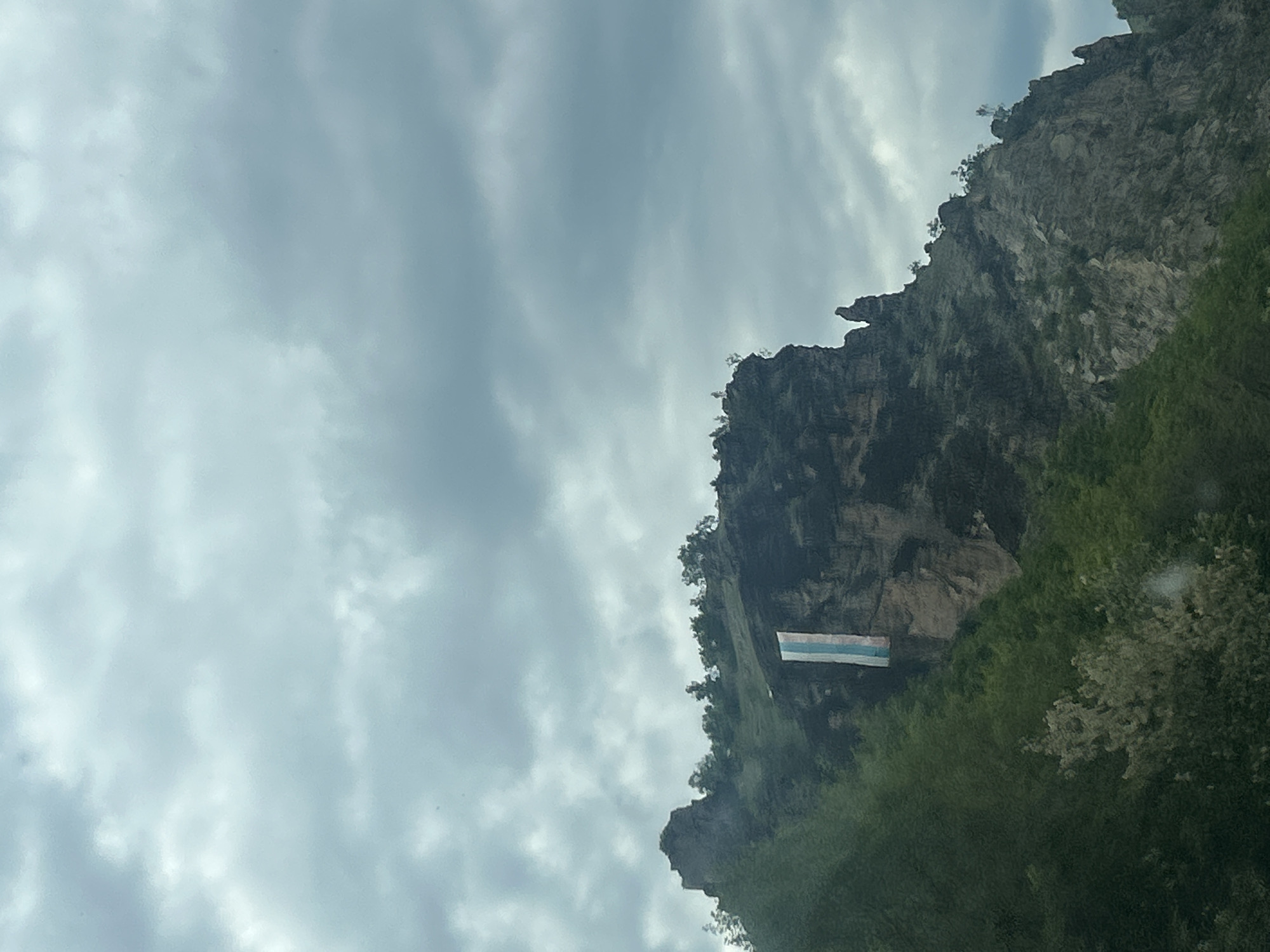 The image shows a view of the rocks and vegetation around Bukovo, Bulgaria. A horizontally oriented rectangle with three stripes, white on top, light blue in the middle, and blue at the bottom, is attached to the rockface. The sky is cloudy.