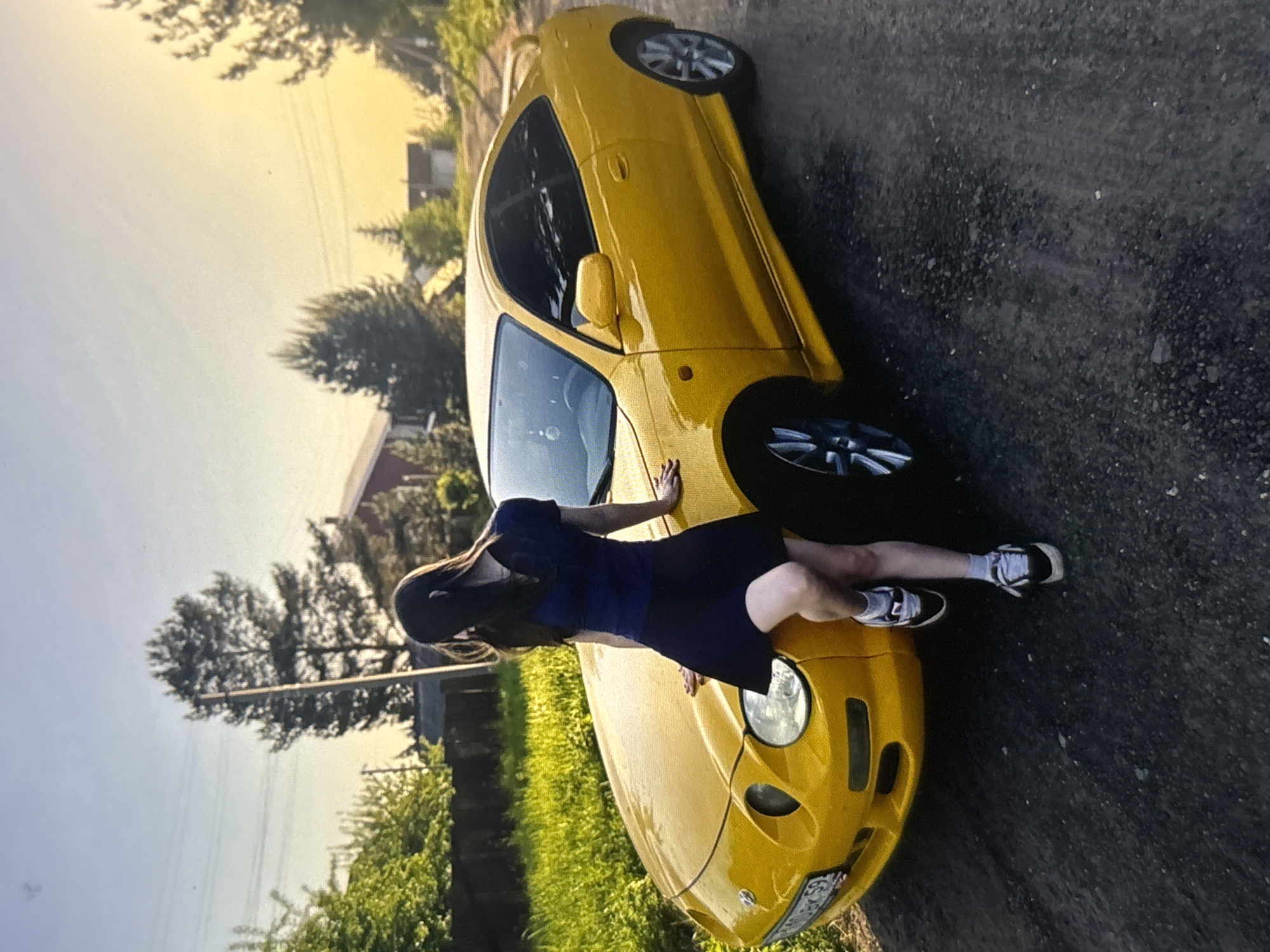 A young woman with dark hair, wearing a dark blue short-sleeved top and a dark blue skirt, stands leaning against the hood of a bright yellow coupe car. Her left leg is bent, with her foot resting on the car's fender, and her right leg is crossed over her left. She is wearing white and black sneakers with white socks. The car, a Toyota Celica, is parked on a gravel and asphalt road. The environment appears to be a rural or suburban area, with trees, grass, and buildings visible in the background under a bright, sunny sky with a golden hue, suggesting either sunrise or sunset. The location is specified as being in Ufa, Russia.