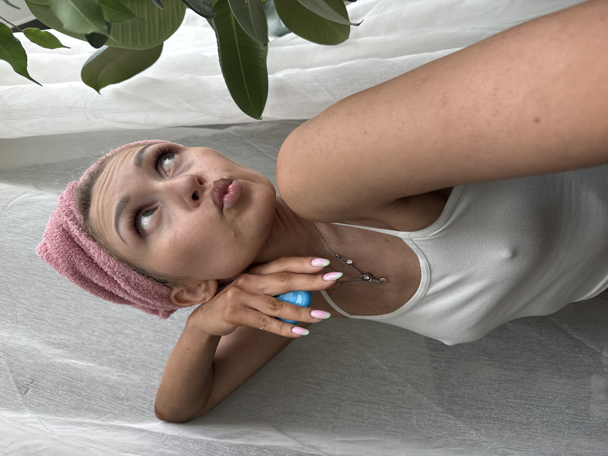 The image presents a young adult female captured in an indoor setting, purportedly located in the city of Chaikovsky, Russia. She is positioned with her right arm bent and resting against a light-colored, vertically draped fabric surface, likely curtains, which forms the background on the left. Her head is tilted, and her gaze is directed upwards and to the right of the frame, with her lips slightly pursed in a contemplative or playful expression. She wears a pink, textured headwrap that covers her hair and a white, ribbed spaghetti-strap tank top, through which the outline of her nipples is visible. Her right hand, adorned with a pink-to-light-green gradient manicure, grasps a small, glossy, ovate blue object. Around her neck, a delicate silver-toned necklace with small charms, including one shaped like a key, is visible. On the far right of the frame, large, dark green leaves of an indoor plant are partially visible, set against the continuation of the white draped fabric. Her skin on the visible arms and shoulder shows light freckling and minor texture.
