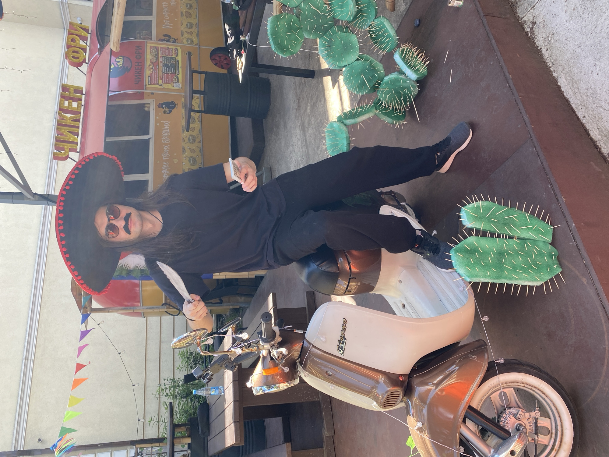 An adult male is prominently featured, seated on a two-tone brown and cream Honda "Steorno" scooter on a raised wooden platform. He has long dark hair, wears dark sunglasses, a fake black mustache, and a large sombrero with a red-trimmed brim. His attire consists of a black t-shirt and black pants with dark sneakers. In his right hand, he holds a serrated utility knife pointed upwards and slightly left. In his left hand, he holds a fanned stack of US $100 banknotes.
The scooter has chrome handlebars, a chrome headlight bezel, and a white-walled front tire, with decorative string lights draped over its front and handlebars. On the wooden platform, two stylized green cacti with toothpick spikes are positioned in front of the scooter, and a larger cluster of similar cacti is to the right of the man's left leg.
In the background, a red and yellow food truck is visible, bearing Cyrillic text "ЧИКЕН ФРИ" (Chicken Fry) on its top sign and side. Menus and chicken illustrations are displayed on the truck's side panels. Behind the food truck is a light-colored building with architectural molding. Above the background elements, colorful triangular bunting and string lights are strung across the scene. To the left, wooden picnic tables and benches are visible, with a plastic water bottle on one table. A black barrel-style table is situated near the food truck, and another dark table is on the raised platform to the right.
The scene is an outdoor setting in Sochi, Russia, under bright daylight, casting distinct shadows. The wooden platform meets a concrete or paved ground surface in the foreground.