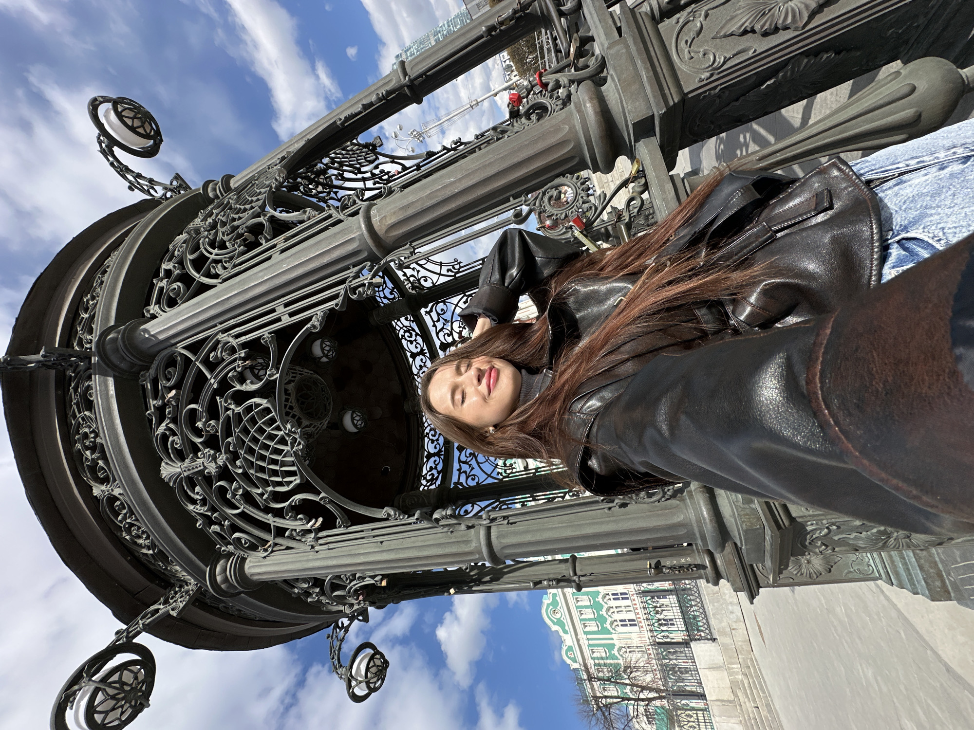 A young woman wearing a black leather jacket and blue jeans is standing under a large, ornate iron archway. The archway has several spherical light fixtures attached to it. The woman has long brown hair and is smiling with her eyes closed. The sky is blue with scattered white clouds. In the background, a green and white building can be seen through the archway. Based on the style of the archway and building, the location is likely in Yekaterinburg, Russia.