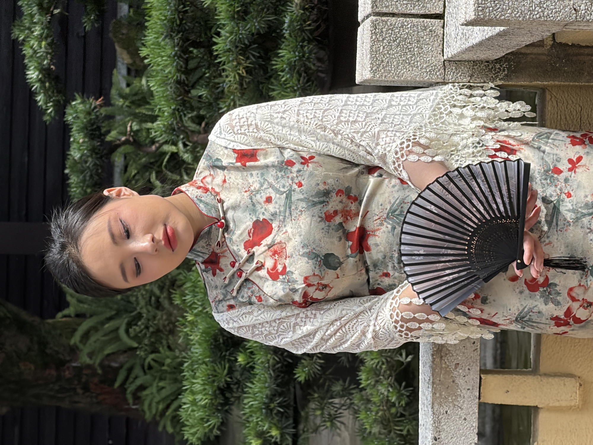 A young East Asian woman stands outdoors, holding a black fan. She is wearing a floral patterned traditional Chinese dress with lace sleeves and red detailing. The background shows lush green foliage and some stone structures. The scene appears to be in a garden or park in Yuzhong District, China.