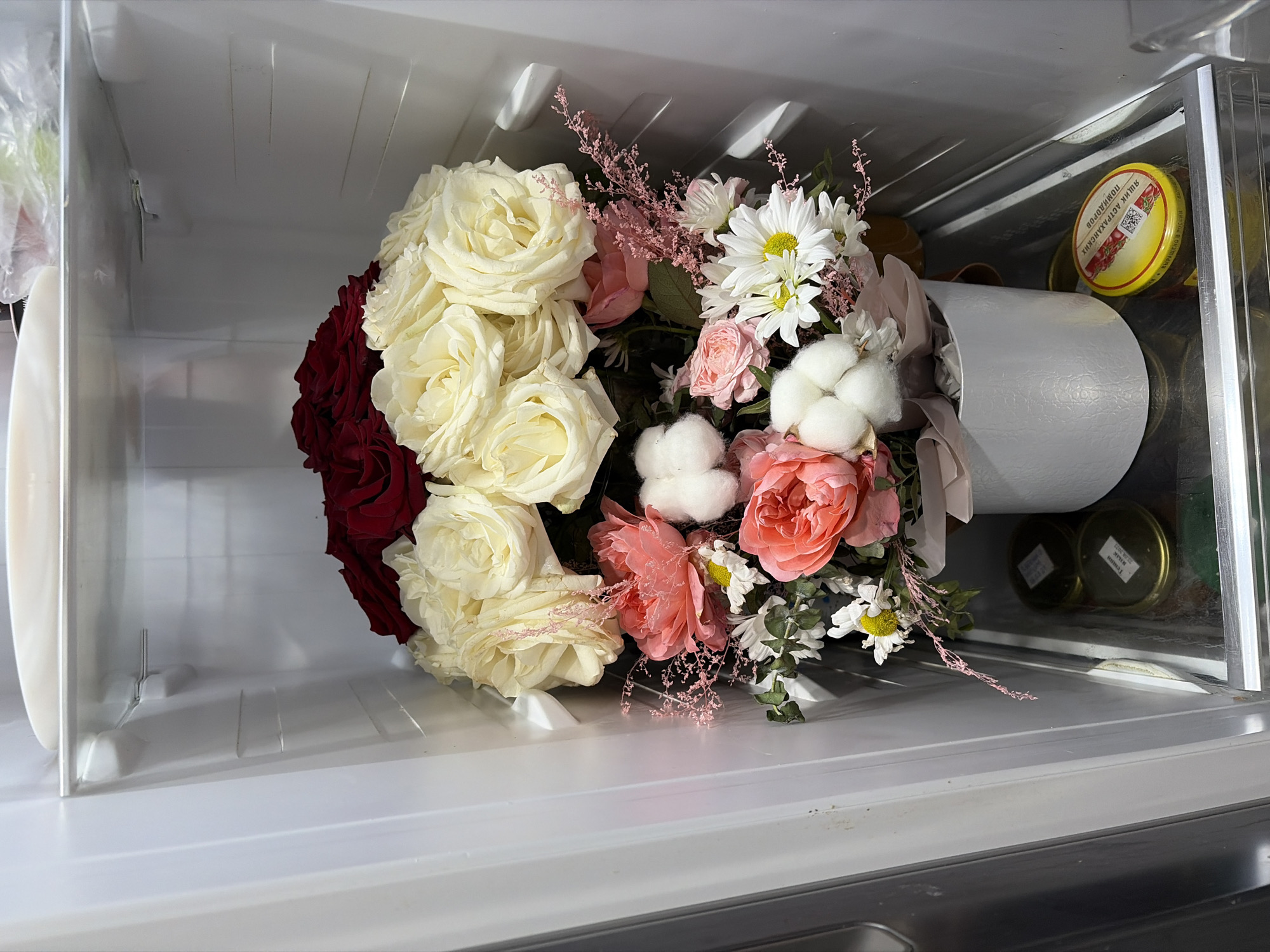 Inside a refrigerator, a bouquet of flowers sits on a shelf. The bouquet is composed of red, white, and pink roses, along with white daisies and cotton bolls. The flowers are arranged in a white container. Several jars are visible on the right side of the shelf. The refrigerator appears to be in a residential or commercial kitchen. This image was taken near the city of Chaikovsky, Russia.