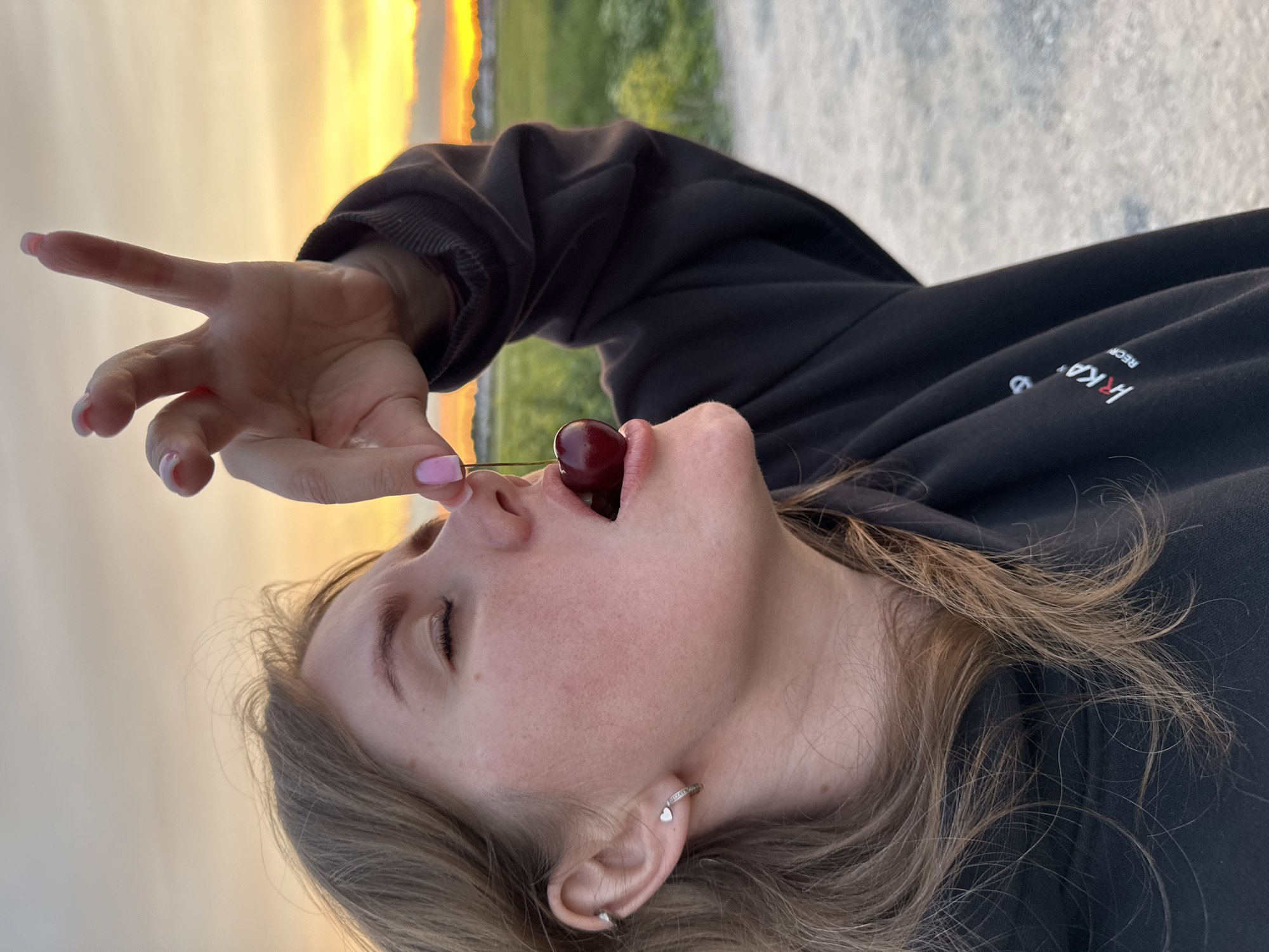 The image depicts a young person, likely female, captured from a low angle and rotated 90 degrees to the left. The subject's head and upper torso are visible, with her face turned upwards and mouth slightly open. A single red cherry, held by its stem between her thumb and forefinger, is positioned directly above her open mouth, suggesting she is about to eat it. Her fingernails are painted a light pink.
Her eyes are closed or mostly closed, conveying a sense of focus or enjoyment in the moment. Light brown, wavy hair frames her face and flows downwards. She is wearing a black hoodie, on which faint white and red text can be seen, partially spelling out "HRKA" and "RECR". Visible on her ear is a small hoop earring with a dangling silver heart charm, and another small stud earring in her earlobe.
The background, also rotated, features a vibrant, warm orange and yellow sky, indicative of either sunrise or sunset. Below the sky, some blurred green foliage and a light-colored ground or path are visible. The setting appears to be outdoors, in a natural or semi-natural environment, potentially in Горный, Russia, during twilight hours. The overall image captures a candid, serene moment of sensory pleasure.