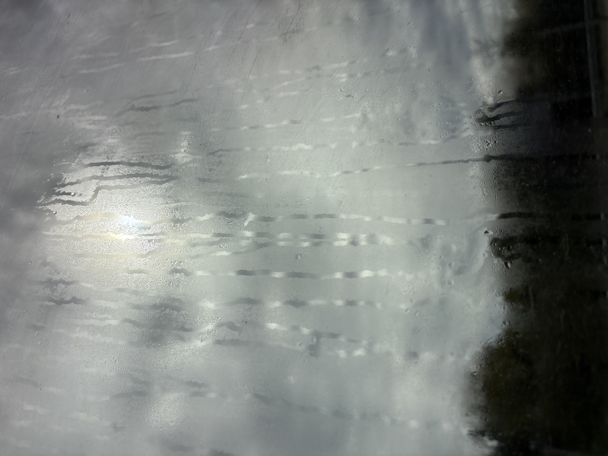 The image shows a view through a wet glass surface. The glass is covered in water droplets and streaks, distorting the scene behind it. In the background, there is a glimpse of the sky with some cloud cover. On the right side, there's a dark, indistinct shape, possibly foliage or a dark structure. The location is not discernible from the image.