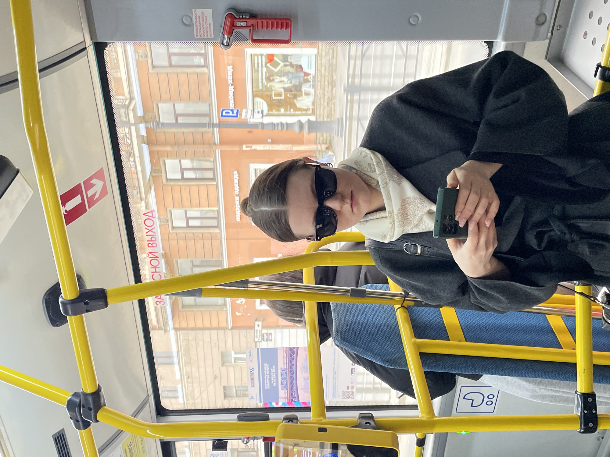 A woman with sunglasses and a dark coat is seen inside a public transportation vehicle (likely a bus or tram) with yellow handrails. She's holding a smartphone in her hands. Outside the window, the buildings of Saint Petersburg, Russia, are visible. The windows include signs with Russian writing. Other passengers are also visible.