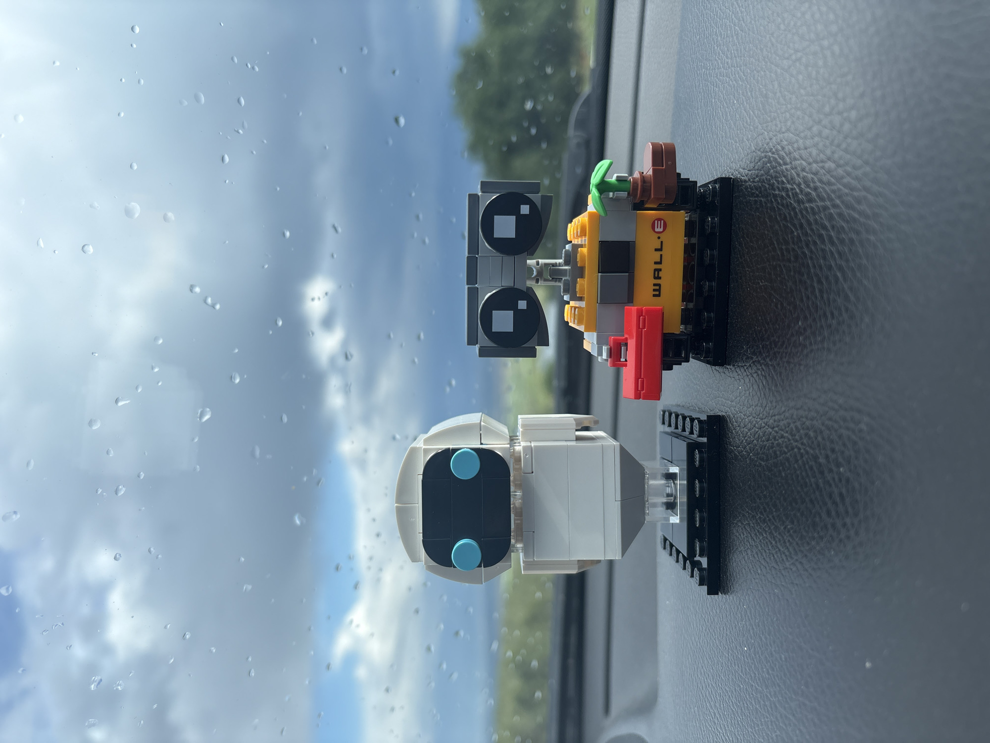 The image shows two Lego figures of WALL-E and EVE positioned on a textured surface, likely the dashboard of a car, judging by the reflection of a cloudy sky and raindrops on glass. WALL-E is constructed with yellow, grey, black, and red Lego bricks and has a plant protruding from its top. EVE is white and grey with blue accents. The location is Unknown, Unknown.