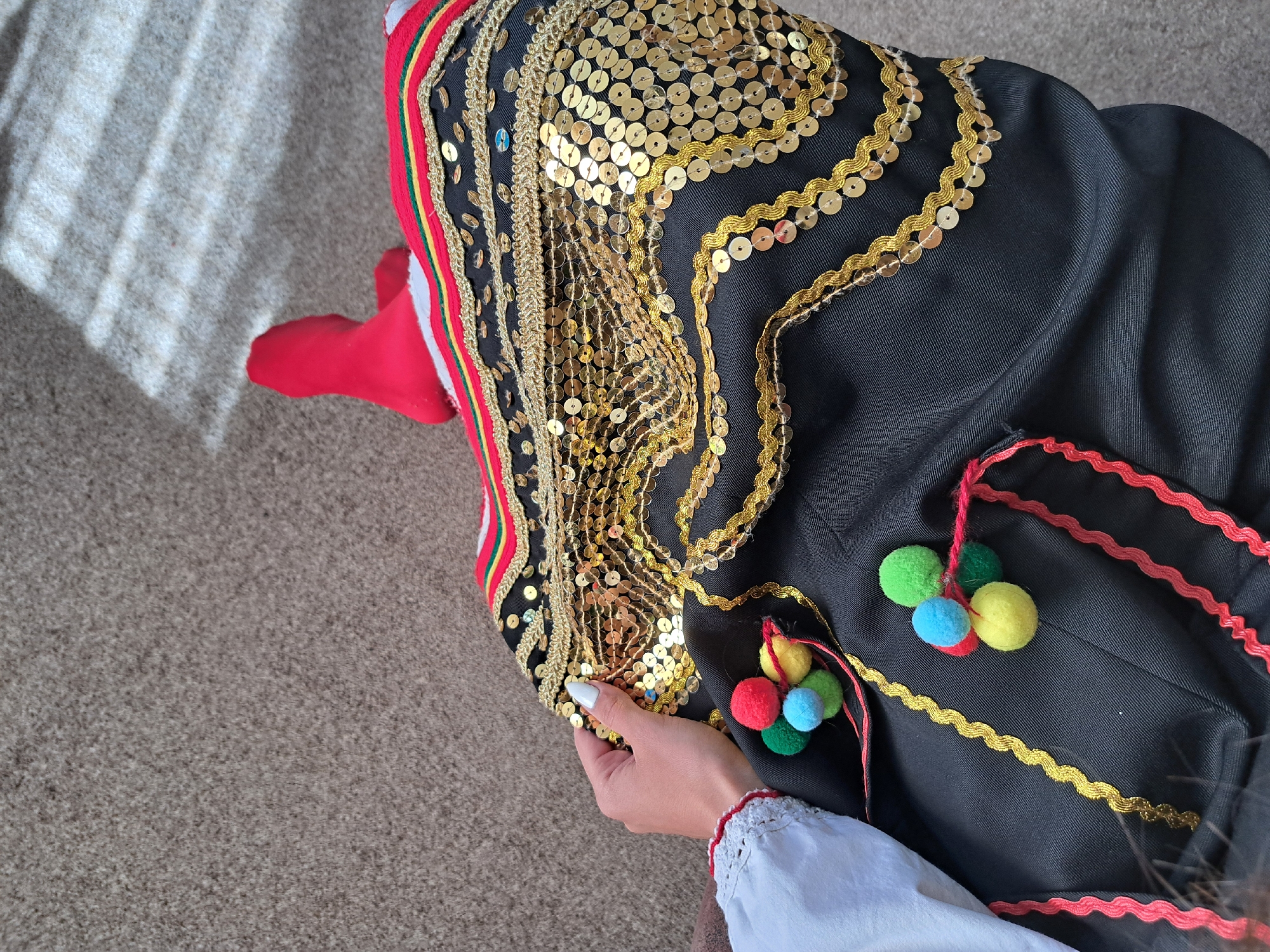 A high-angle shot shows a person holding a traditional costume on a carpeted surface. The costume features a black base adorned with multiple rows of gold sequins and gold trim. Additional embellishments include a red, yellow, and green stripe on one edge, along with colorful pom-poms attached by red strings. The person holding the costume is wearing a white garment with a red trim detail at the wrist. The location is in the city of Unknown, Unknown.