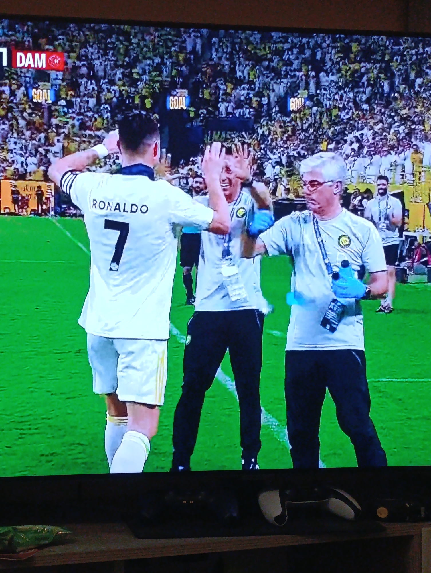 The image shows a soccer (football) game. Cristiano Ronaldo, wearing a white jersey with the number 7 and the name "RONALDO" on the back, is high-fiving two men in similar light-colored shirts and black pants. The men appear to be part of the team's support staff. One of them is wearing blue gloves and holding water bottles. In the background, there is a large crowd of spectators, most wearing white and yellow clothing. Signs with the word "GOAL" are visible. The scoreboard shows "DAM" with a 1 score. The location is not discernible from the image itself. Plovdiv, Bulgaria is not identified within the image. An Xbox controller is visible at the bottom of the image, suggesting the photo was taken from a TV screen.