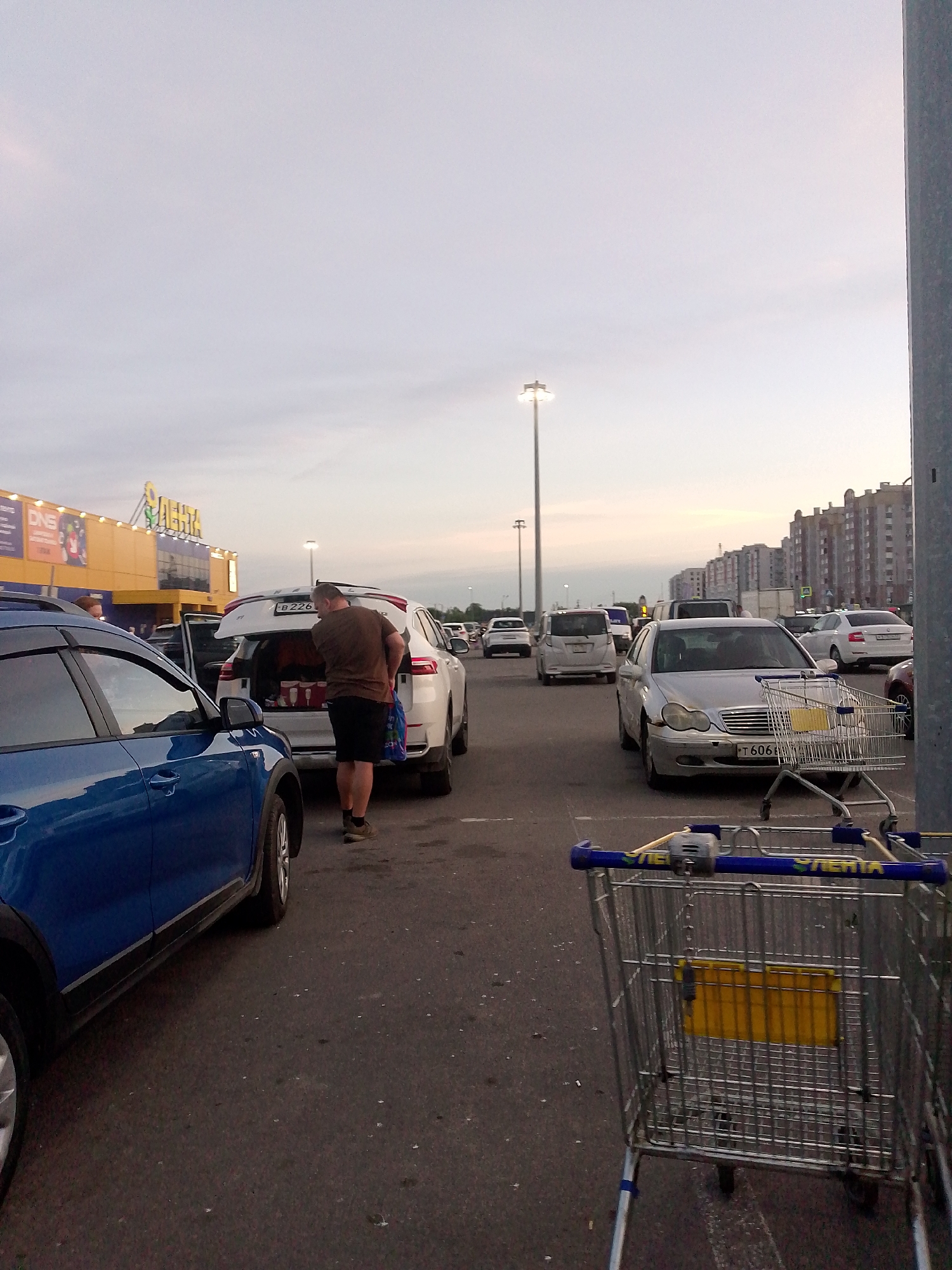 In the city of Shimsk, Russia, a man stands by the open trunk of a white SUV, placing items inside. The SUV's license plate reads "B 226". In the foreground, a blue car is parked, facing away from the viewer. To the right of the SUV, a silver sedan with license plate "T 606 B" is also parked. A shopping cart with a yellow insert is positioned in front of the silver sedan. In the background, other cars are visible in a parking lot, and a large yellow building with signage is present. The sky is overcast with a pale hue.