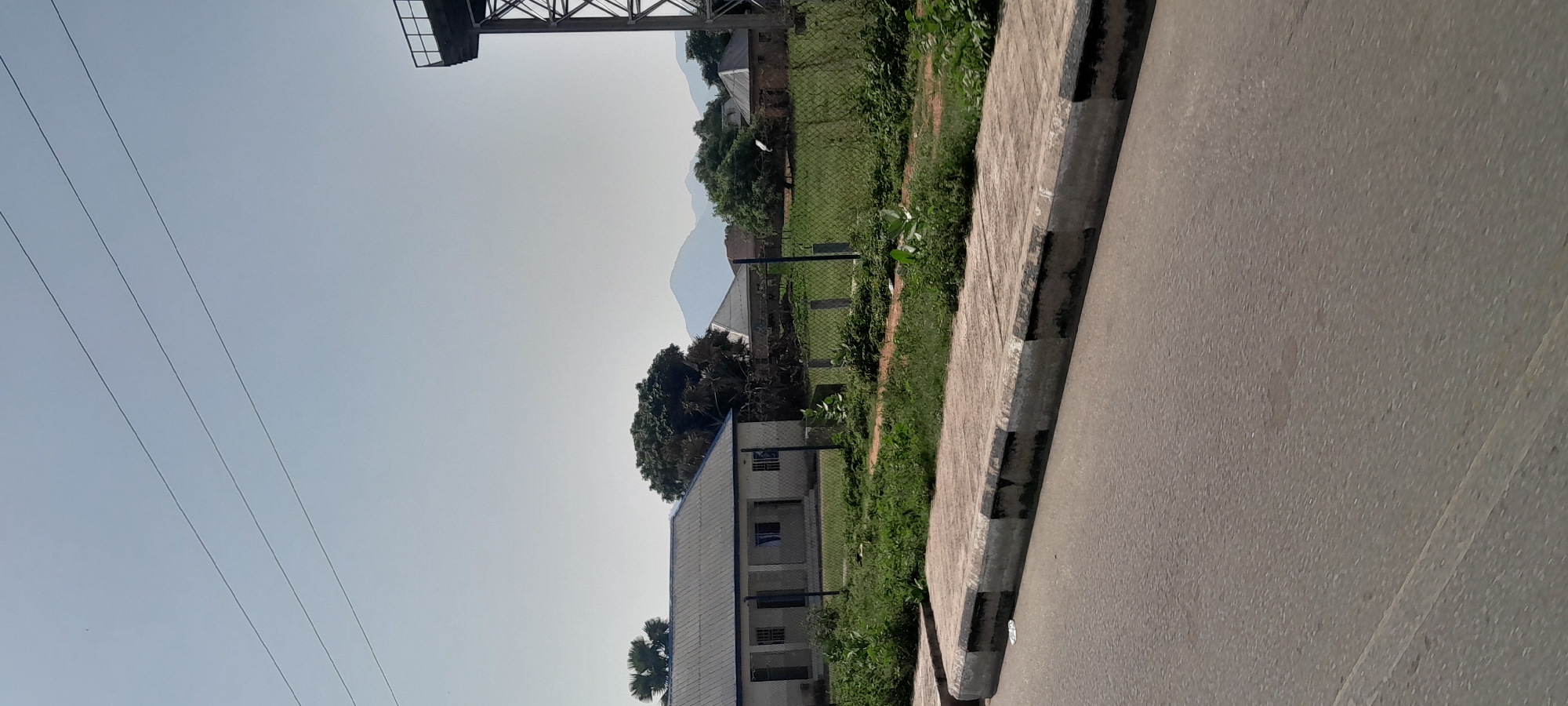 The image shows a street scene in Jalingo, Nigeria. In the foreground, a paved sidewalk with black and white striped edging runs along a road. Beyond the sidewalk is a grassy area enclosed by a chain-link fence. Several buildings, likely part of an institution or complex, are visible behind the fence. These buildings appear to be single or two-story structures with light-colored walls and darker roofs. Some trees, including palm trees, are interspersed within the property. In the background, hazy mountains or hills can be seen, suggesting a rural or semi-rural setting. The sky is clear and bright, indicating daytime, likely morning or afternoon. The image is captured from a low angle, close to the ground, and is oriented vertically. There are no people visible in the image, and no text is discernible. The overall impression is one of a quiet, functional area under a clear sky.