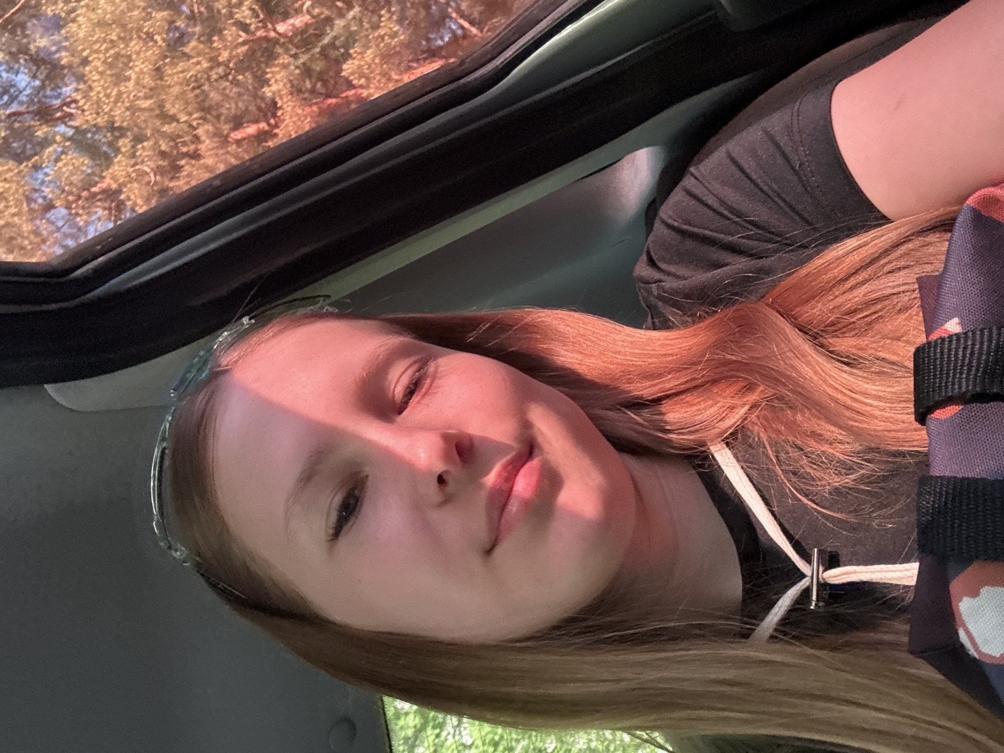 A young woman is photographed from a low angle within a car, likely sitting in the passenger seat. She has fair skin, light brown hair with reddish highlights, and is wearing a dark-colored shirt. A pair of decorative glasses rests on her head. Part of a dark-colored bag or object is visible near her shoulder. Outside the car window, tree branches are visible, suggesting a location with trees nearby, possibly a wooded area or park. The angle suggests the photo was taken while looking up through the car's window or sunroof. No specific details indicate the location as Chaikovsky, Russia.
