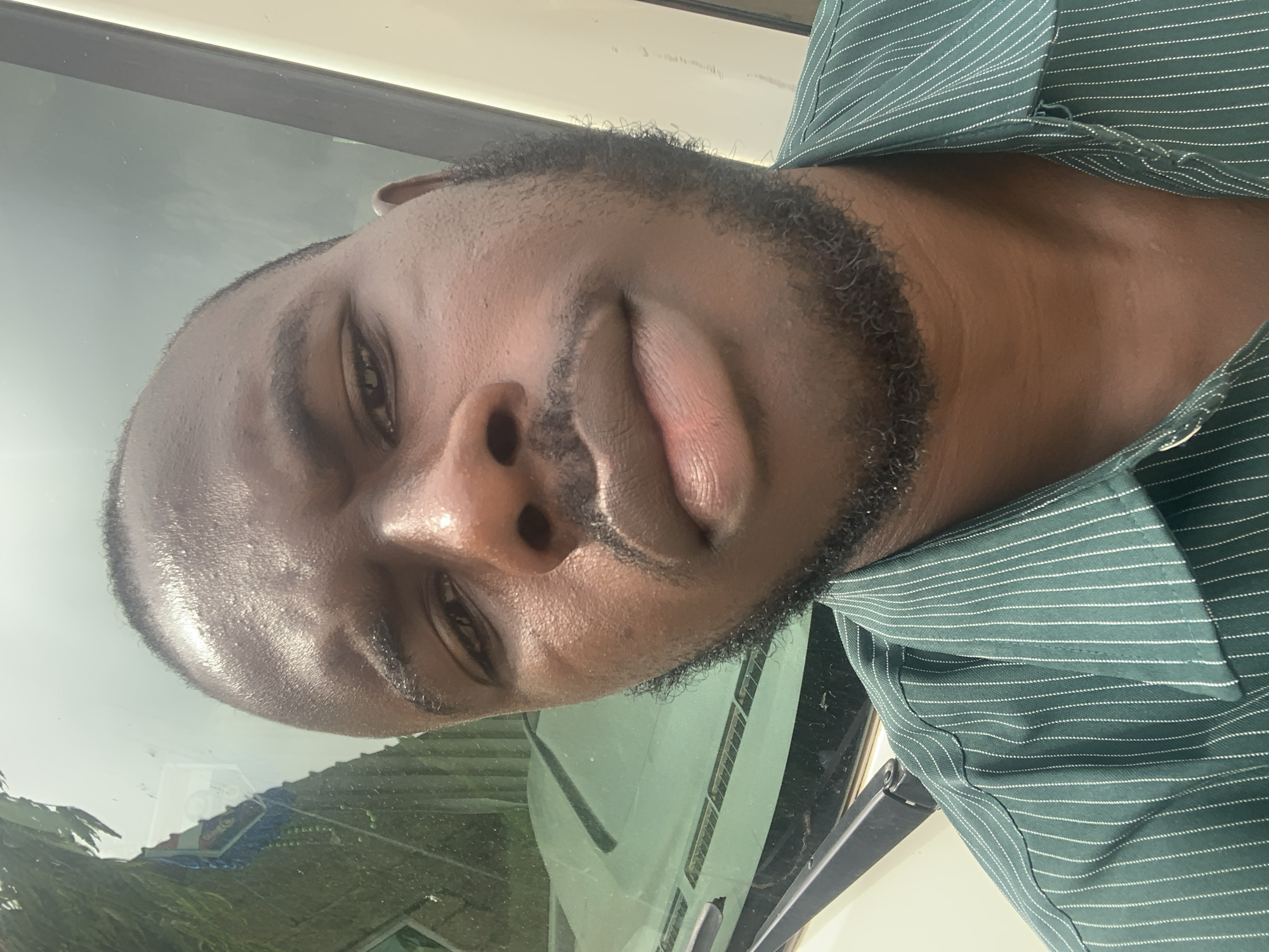 A close-up self-portrait photograph shows a person with dark skin and short, dark hair and a beard, wearing a green pinstriped shirt. The person is gazing directly at the camera. A car windshield and building are visible in the background. The location is potentially Jos, Nigeria. The exact location is unclear.