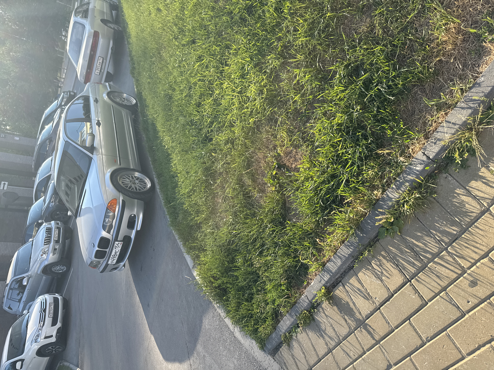 The image displays a row of parked cars along a grassy verge next to a sidewalk made of interlocking bricks. The scene appears to be a residential or commercial area in Koltsovo, Russia, given the license plates. The time of day is late afternoon, indicated by the long shadows cast by the grass and trees onto the sidewalk. The weather seems pleasant, with bright sunlight illuminating the scene.
The primary objects are several cars, including a silver BMW, a silver Toyota Land Cruiser Prado, and a white Citroën. The BMW has a license plate that reads "P498MC 154". The Toyota Land Cruiser Prado has a license plate "E921CP 154". The Citroën's license plate is partially visible as "X377OY 154". The cars are parked on an asphalt surface.
The setting is outdoors, with a neatly manicured lawn bordering a paved walkway. The perspective is from a slightly elevated angle, looking down at the cars and the surroundings. No people are visible in the frame, and there are no obvious signs of any specific event taking place; it appears to be a typical day with cars parked. The overall mood is peaceful and ordinary.