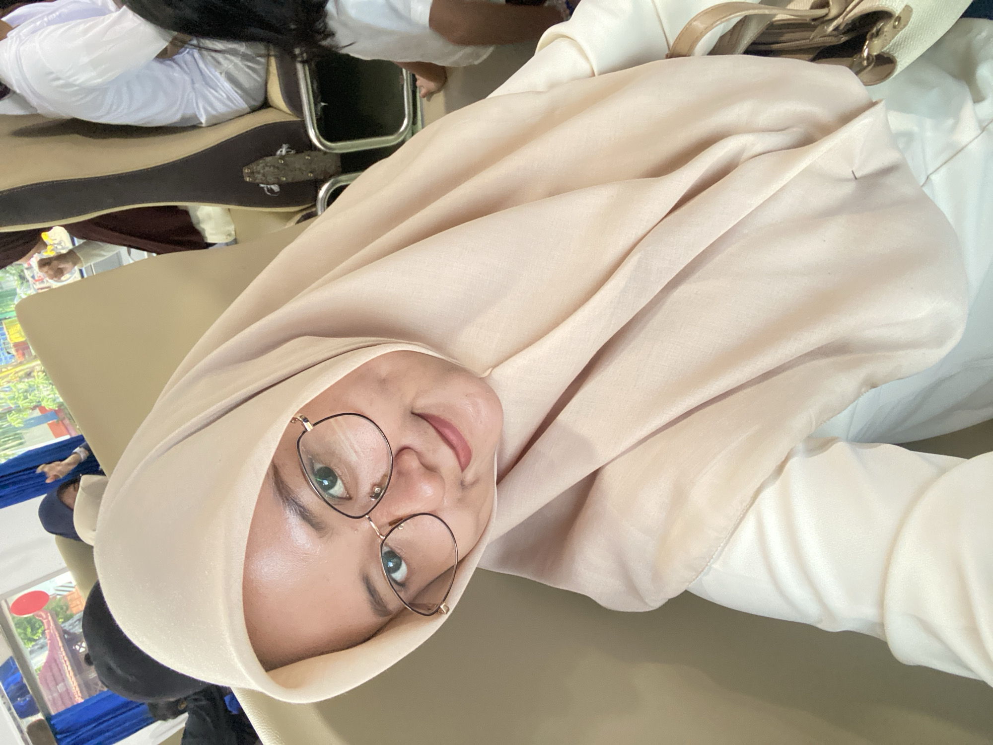 A woman wearing a beige hijab and round glasses is the focal point. She is looking at the camera with a slight smile. She's indoors, possibly in transit, as suggested by glimpses of other people and scenery through windows behind her. The location may be Probolinggo, Indonesia, but this is not visually confirmed in the image itself.