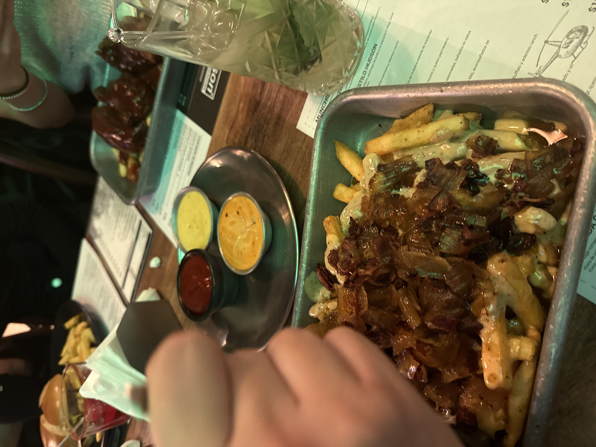 This image depicts a casual dining scene at a restaurant or bar in San Andrés Cholula, Mexico, likely in the evening given the dim, artificial lighting that casts a greenish tint. The primary subject is a metal tray overflowing with loaded French fries, generously topped with a creamy white sauce (possibly cheese or a savory dressing) and a large portion of what appears to be caramelized onions or crispy bacon bits.
Next to the fries, a small, circular metal plate holds three dipping sauces in individual cups: bright red (ketchup), a vibrant yellow (likely mustard), and another orange-yellow sauce. In the upper left background, another metal tray contains what seem to be BBQ ribs, coated in a dark, glossy sauce. A glass with a textured surface, containing a beverage garnished with mint leaves, is also visible.
A person's hand is prominently in the foreground, seemingly holding a camera or phone, while another hand with a bracelet is visible in the background on the left. All items rest on a wooden table, with menus or paper placemats visible underneath the dishes.
Visible text from the menus includes "MUNCHIE" and "STILO HUDSON". Food descriptions mention "tociño y queso parmesano" (bacon and parmesan cheese), items "sazonadas, bañados en salsa bbq, acompañados de" (seasoned, bathed in BBQ sauce, accompanied by), and "buffalo y aderezo ranch" (buffalo and ranch dressing), along with "dijon" and "queso". A partial price of "$1$" is also visible.