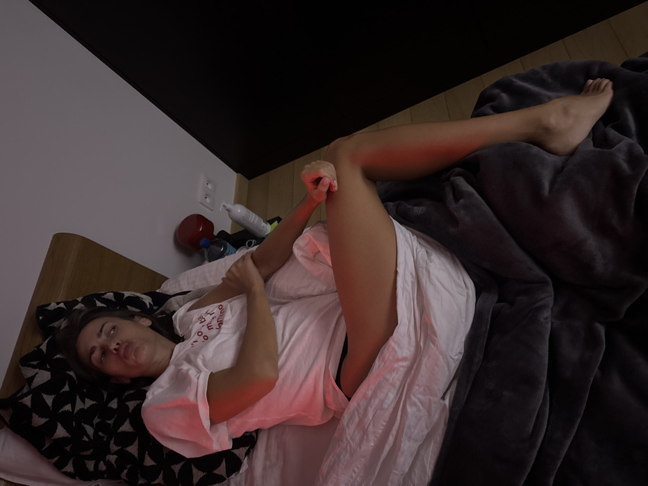 A woman lies in bed with her leg extended towards the camera. She is partially covered by a white sheet and a dark gray blanket. Her head rests on a black and white patterned pillow. Various items, including a red container and a bottle, are visible on the bedside table. The room has a combination of white and dark walls.