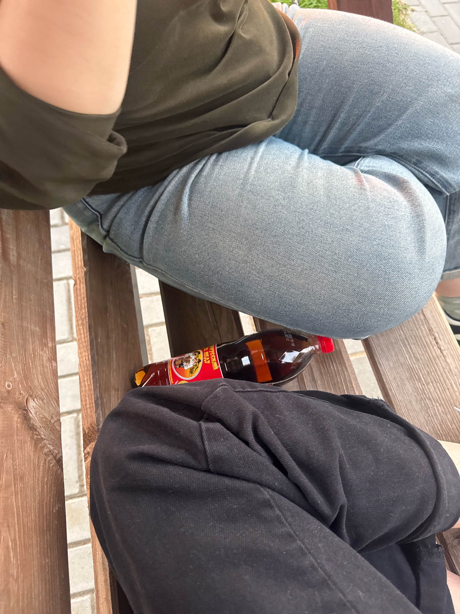 The image shows a close-up view of two individuals seated on a wooden bench. One person is wearing a dark green long-sleeved shirt and light blue jeans, while the other is wearing a black hooded sweatshirt and black pants. A bottle of dark liquid with a red cap, possibly a beverage or sauce, rests on the bench between them. The background reveals a paved area with white tiles, suggesting an outdoor public space. The image does not contain any identifiable landmarks or features that would definitively place it in the city of Myadzyel, Belarus.