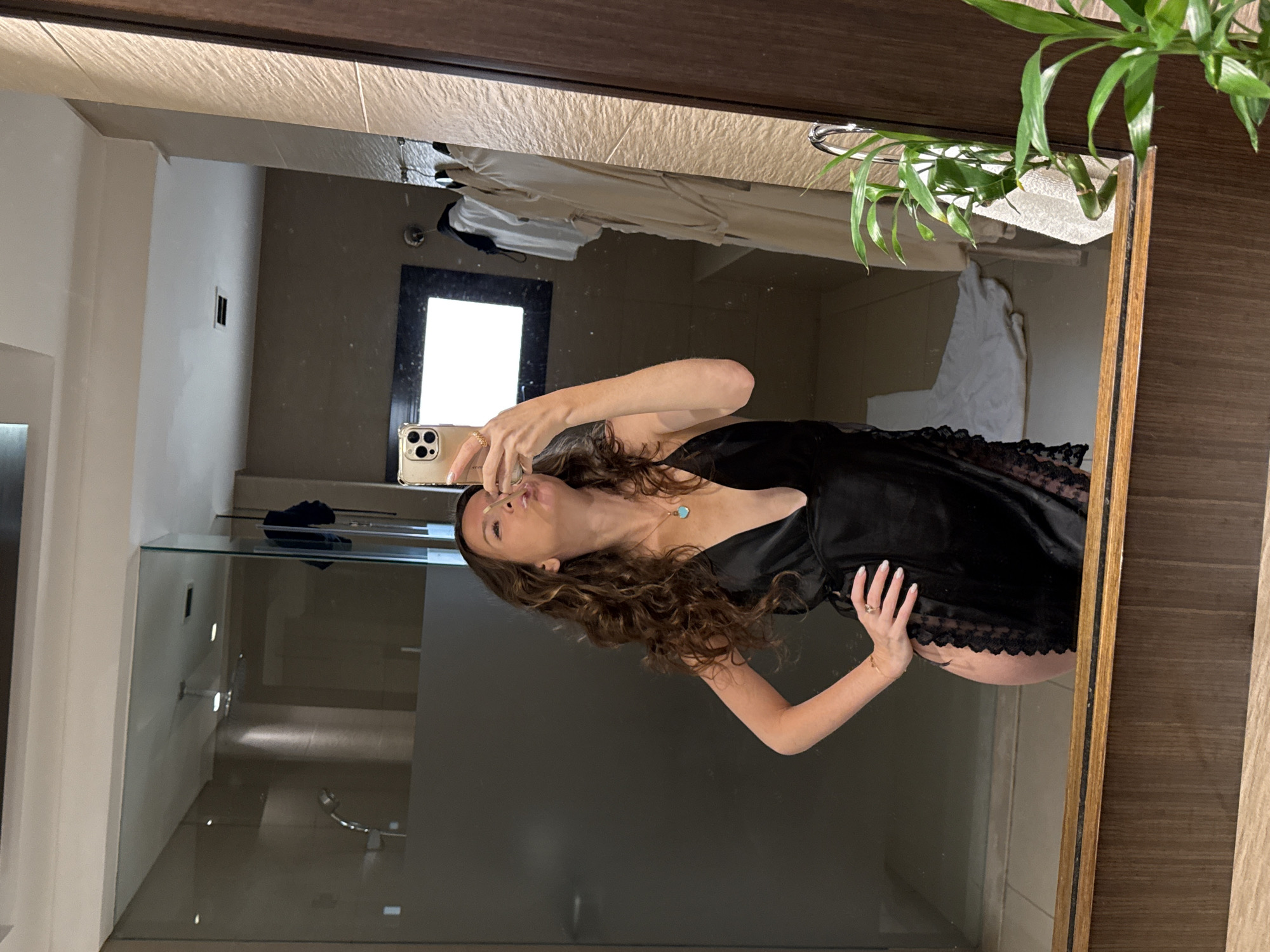A woman in a black slip dress is taking a selfie in a bathroom mirror. She is wearing a necklace with a blue pendant and rings on her fingers. The bathroom appears to be in a hotel, with robes hanging on a rack and a white towel on the floor. The room is modern and well-lit. There are plants in the foreground, and the overall impression is one of luxury and relaxation.
