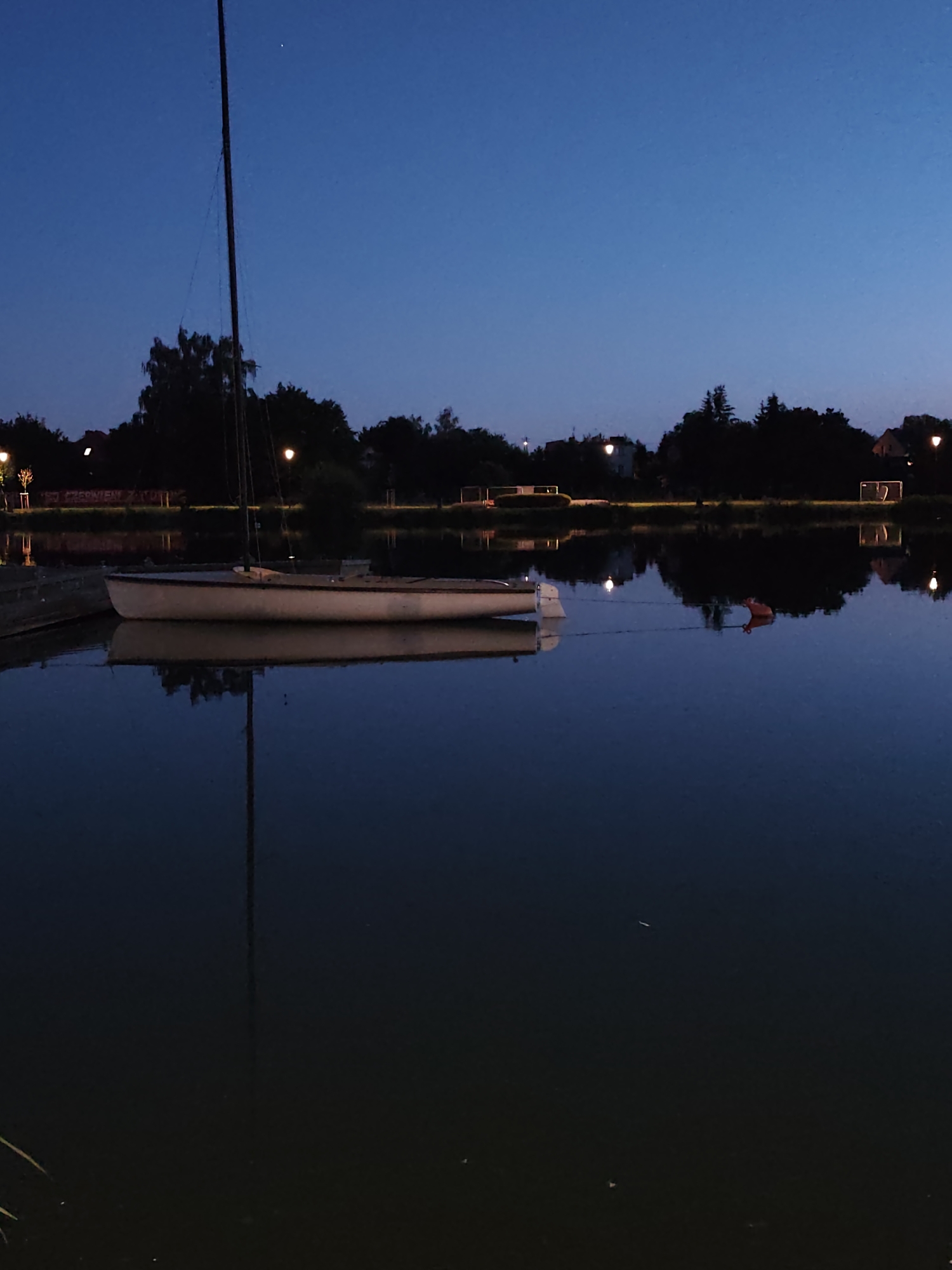 A sailboat is docked on a calm body of water during twilight. The boat, light in color, has a mast that extends high into the dark blue sky. The sky is clear with a faint hint of stars visible. In the background, silhouetted trees and buildings line the shore, with streetlights casting a warm glow. The water perfectly reflects the scene above, creating a mirror image of the boat, mast, trees, and lights. An orange buoy floats on the water to the right of the sailboat. The overall mood of the image is peaceful and serene.