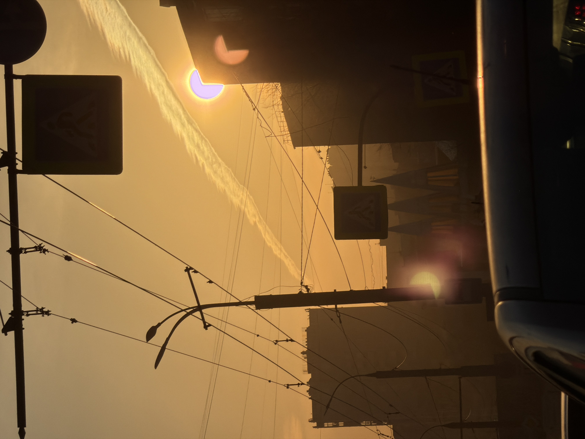 The image depicts a street scene in Izhevsk, Russia at sunset. The sun is partially obscured by buildings, creating a strong glare and silhouetting objects. A contrail cuts across the sky. Power lines and streetlights crisscross the sky. There are street signs visible on poles. A portion of a silver vehicle is on the right of the frame. The overall tone is hazy and warm due to the setting sun.