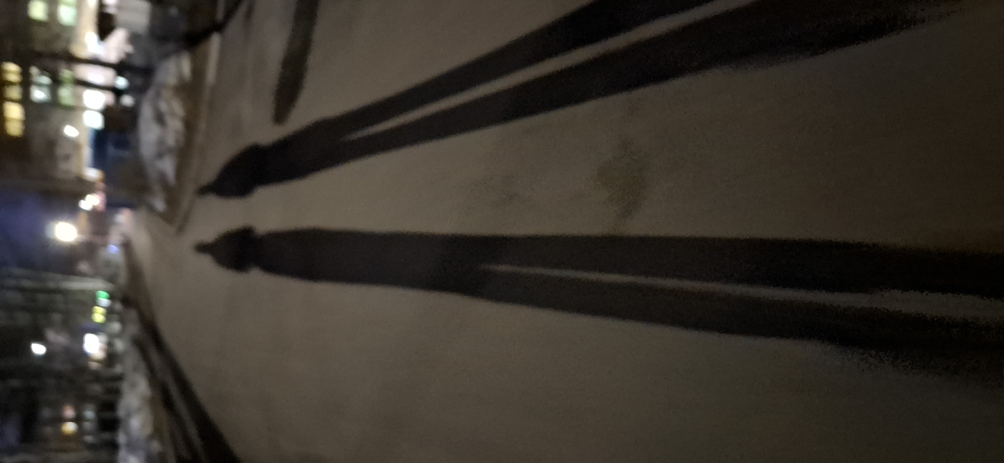 The image shows a dark, blurry street scene at night in what is likely Chaikovsky, Russia. The focus is on a light-colored, possibly concrete, wall or surface that dominates the right side of the image. Two dark, parallel lines run across the wall at an angle. On the left side of the image, indistinct lights and shapes suggest buildings and streetlights along a street. The overall scene is dim and unclear.