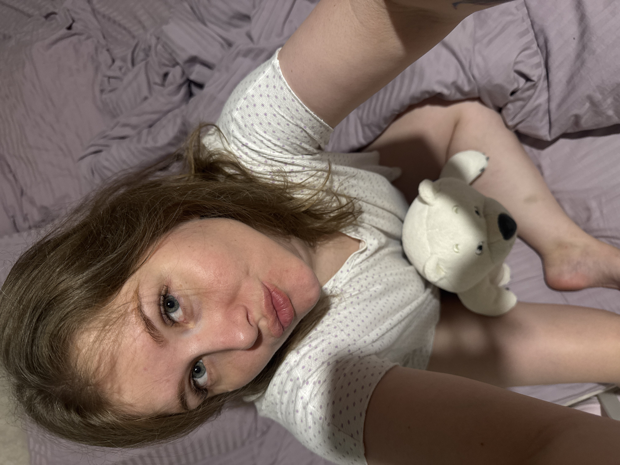 A young woman is captured in a high-angle selfie, lying on what appears to be a bed covered with light purple, subtly striped or textured bedding. Her head is angled upwards towards the camera, with her brown, shoulder-length hair fanned out around her. She has striking blue eyes that gaze directly at the lens, and her lips are slightly pouted, conveying a thoughtful or playful expression. She is wearing a light-colored t-shirt adorned with small purple polka dots.
Next to her, positioned on the bed near her legs, is a small, white polar bear plush toy. The setting appears to be indoors, likely a bedroom, characterized by the soft bedding. The lighting is diffused, suggesting an indoor environment without strong natural light sources. The overall scene implies a relaxed and personal moment. No text is visible in the image. The photo was taken in Gornyi (Dvurechenskoe S/P), Russia.