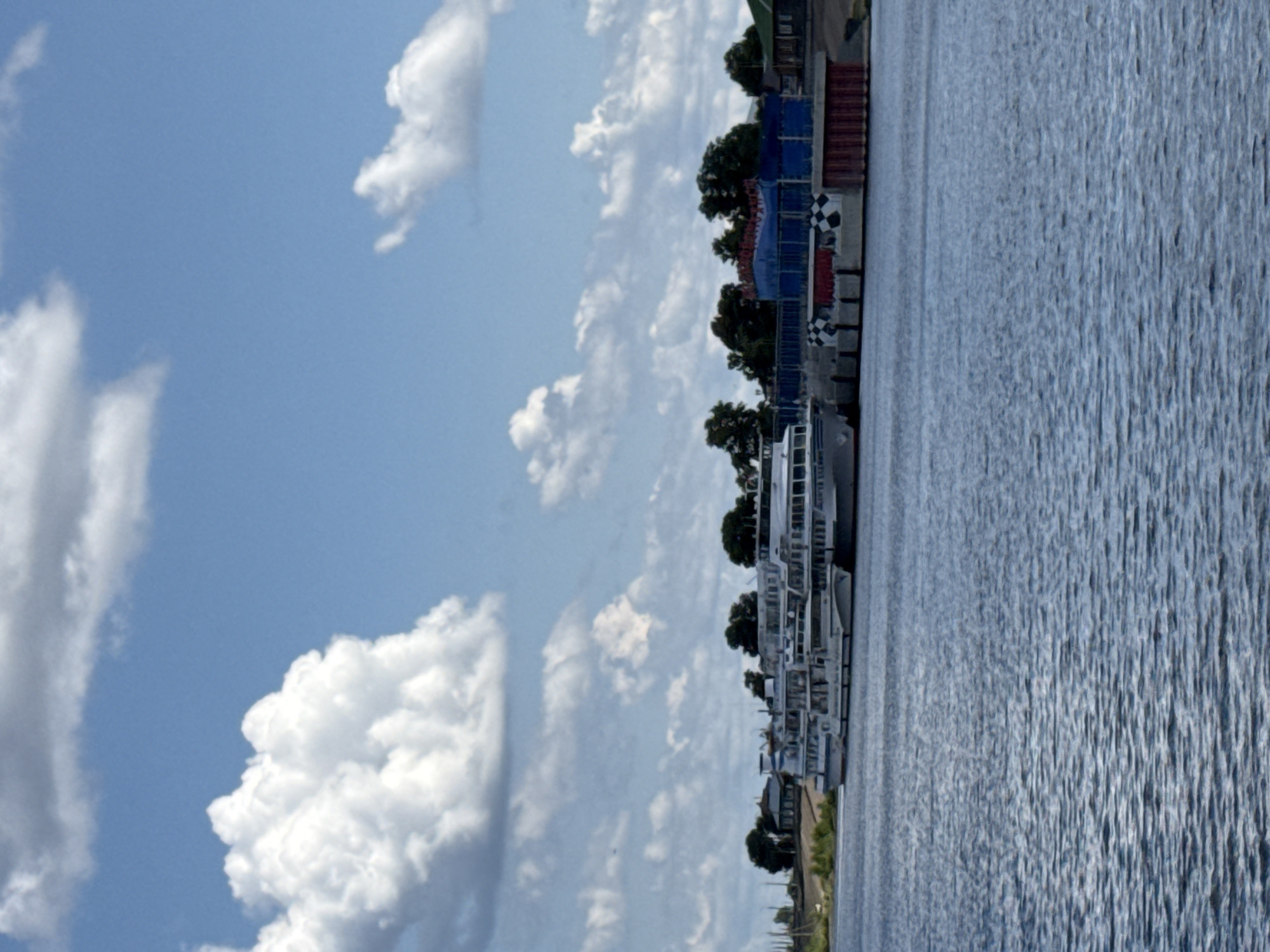 This is a vertical, medium-shot, eye-level photograph of a wide, calm river with a fleet of white passenger ships docked at the embankment on the far side. The sky is bright blue with numerous large, fluffy white clouds. The river occupies the lower half of the frame and is rendered with a choppy texture of dark blue ripples. The embankment on the far side features lush green vegetation in the foreground, behind which are several small, distant houses and buildings, some with dark roofs and green walls. A cluster of dense, dark green trees forms a backdrop to the buildings.
The ships are docked side-by-side, and their white hulls and multiple decks are visible. The lead ship, closest to the viewer's left, appears to be a longer vessel, possibly a motor ship. Behind it are several other similar ships, lined up along the quay. In the center of the docked ships, there is a brightly colored structure constructed of blue and red panels, with some red-colored fencing or netting along the top. This structure appears to be a temporary installation or a building at the port. The checkered black and white patterns on the quay in front of the colorful structure suggest a visual element or marking.
The overall atmosphere is peaceful and summery. The image depicts a serene river scene in the city of Kozmodemyansk, Russia, a location known for its Volga River port. There are no people visible in the image. The scene is static, with the ships at rest and the water smooth.