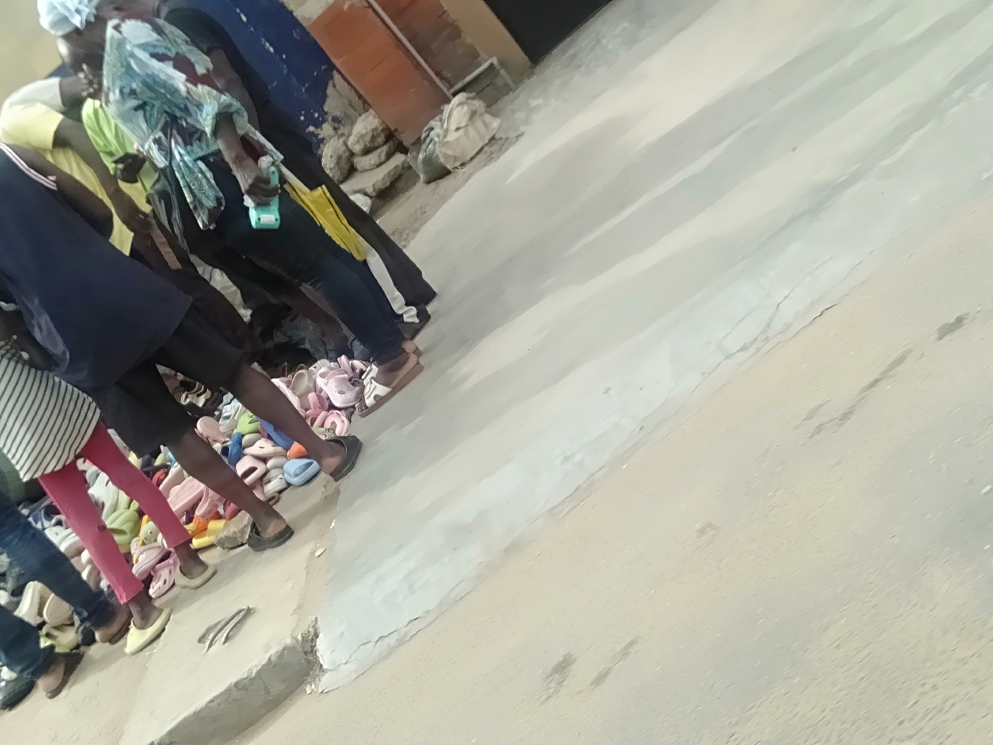 In Shangisha, Nigeria, a group of people stand beside a large collection of shoes. They appear to be browsing or selling. The area has a concrete surface and adjacent structures.