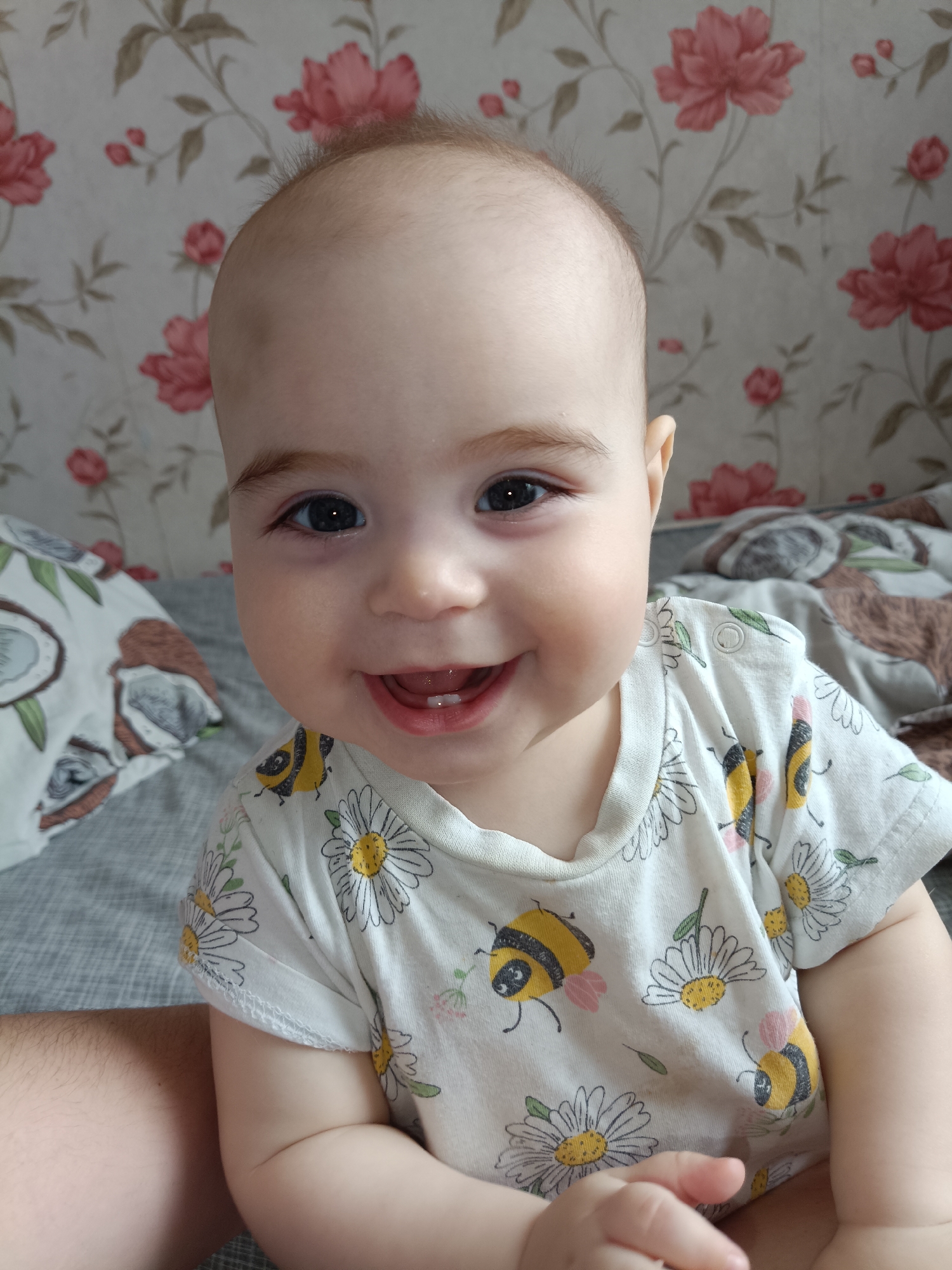 A baby is smiling at the camera. The baby has light skin, dark eyes, and is wearing a white shirt with daisies and bees printed on it. The baby is sitting on a surface covered with a patterned sheet or blanket. In the background, there is a wall with a floral pattern. The location is Noviy, Russia.