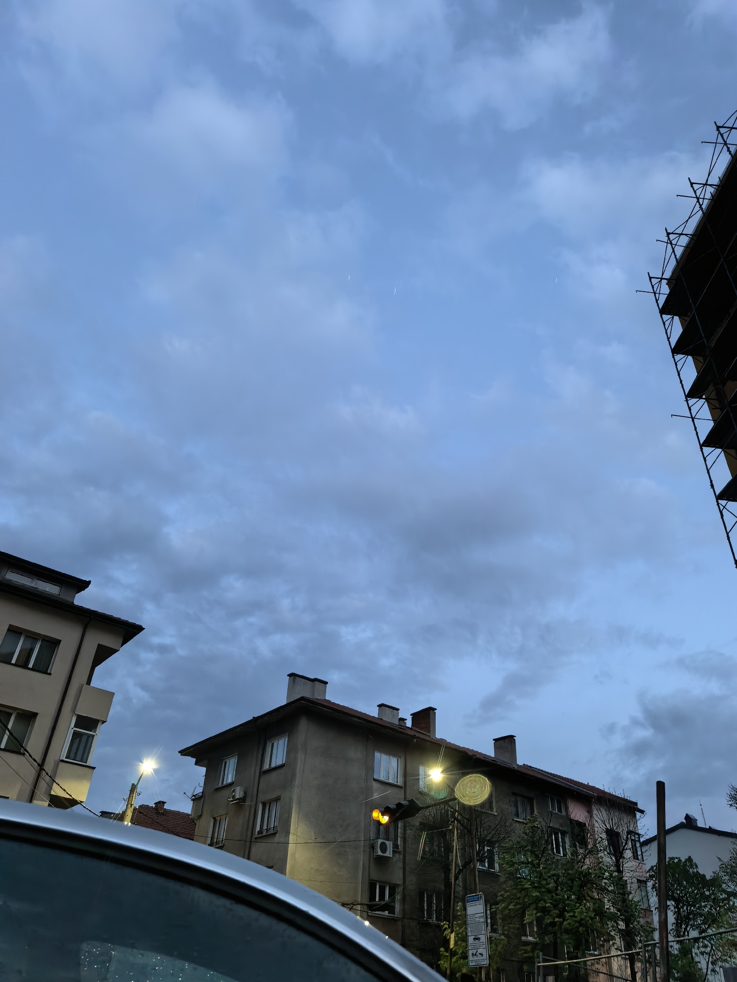 An urban street scene captured during dusk or early evening, characterized by a dark blue-grey overcast sky with scattered clouds. In the upper sky, faint, thin white lines are visible, possibly aircraft contrails. The composition looks upwards, revealing parts of several buildings and construction scaffolding against the sky.
On the left, a portion of a light-colored building with a sloped roof and windows is visible. In the center, a multi-story building with a grey facade, several windows, and multiple chimneys on its roof stands. A bright street light, emitting a star-like flare, is prominent in front of this building, casting a strong illumination. Below it, a yellow traffic light or signal is also visible. Trees and bushes partially obscure the lower part of the central building.
To the right, a large section of dark scaffolding indicates ongoing construction or renovation, silhouetted against the dim sky. A small part of another building is barely visible behind the scaffolding. In the immediate foreground, the metallic silver roof and windshield of a car are visible in the lower left, reflecting some light. The overall mood is quiet and muted, with artificial lights beginning to dominate as natural light fades. A white sign on a pole near the central street light is present, but any text on it is unreadable.