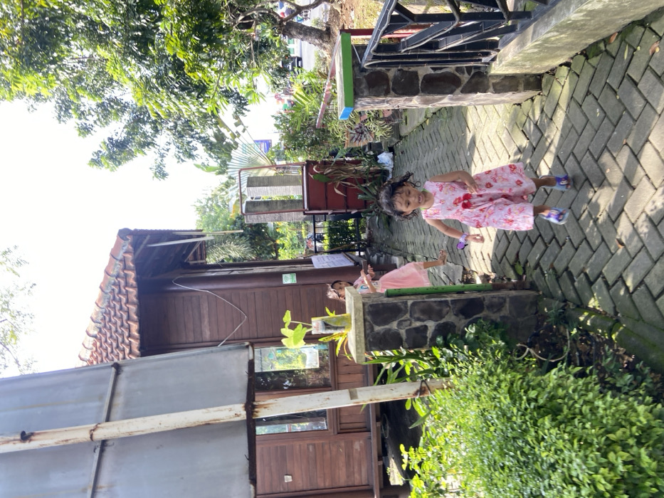The scene is located in the city of Kecamatan Sidoarjo, Indonesia, at the coordinates -7.441 latitude and 112.705 longitude. A young girl in a pink floral dress walks down a brick pathway, smiling. Another young girl in a pink dress walks behind her, also smiling. The girls are walking on a pathway next to a wooden building with a red tile roof. The pathway is lined with bushes and trees, and a wooden fence is on the right. The day is sunny and the scene is peaceful.