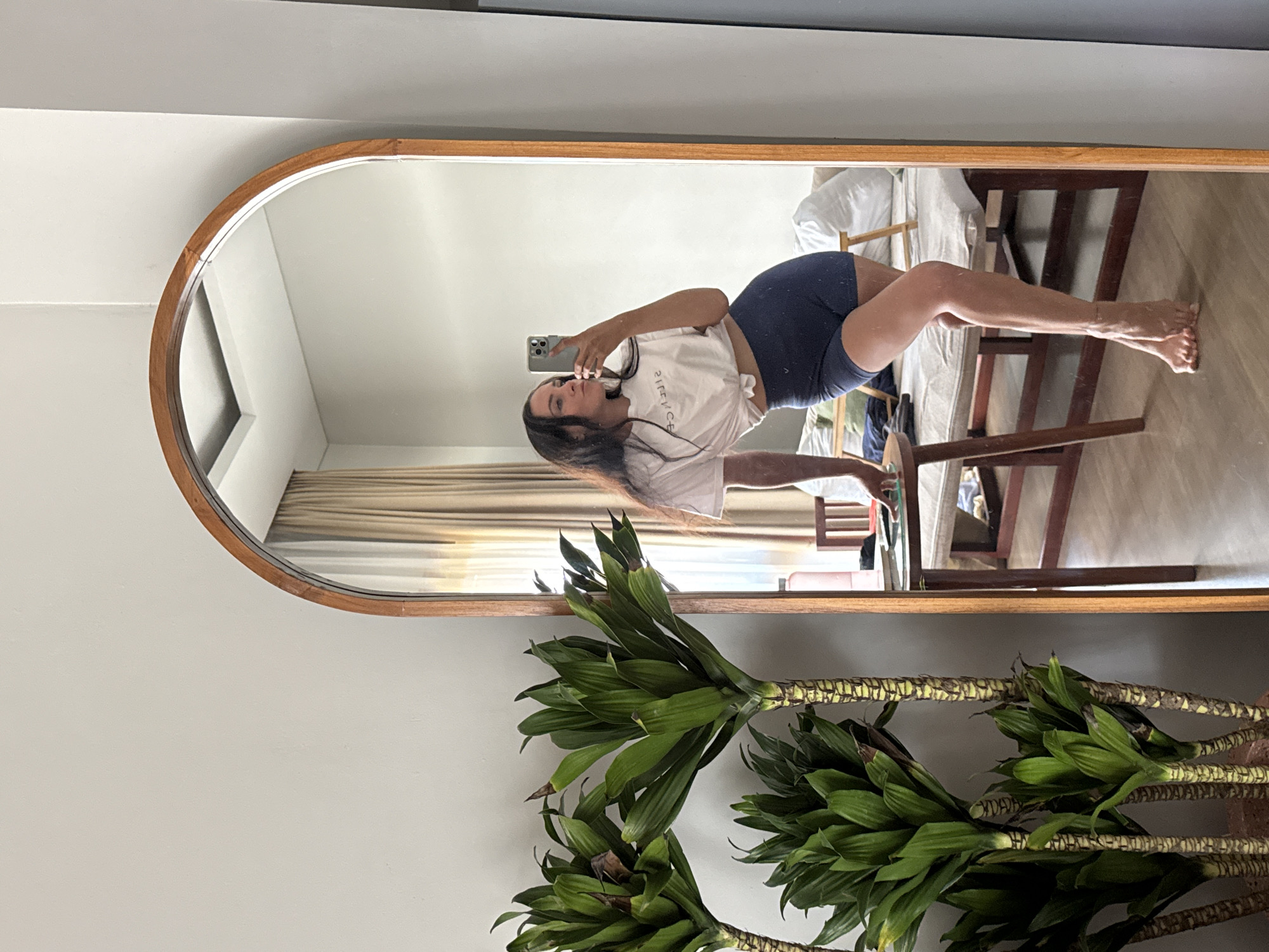 In North Kuta, Indonesia, a young woman with long, dark hair stands by a tall, arched mirror, taking a selfie with her phone. She wears a white t-shirt knotted at her waist and dark blue shorts. Her left arm is extended, holding the phone, while her right hand rests on the mirror's wooden frame. She is positioned leaning forward, with her legs crossed and her right foot raised on its toes. Behind her, visible in the mirror's reflection, is a room with beige curtains, a bed, a wooden chair, and a small table. In the foreground, to the left of the mirror, are three large potted plants with broad green leaves and textured stalks. The lighting suggests it is daytime.