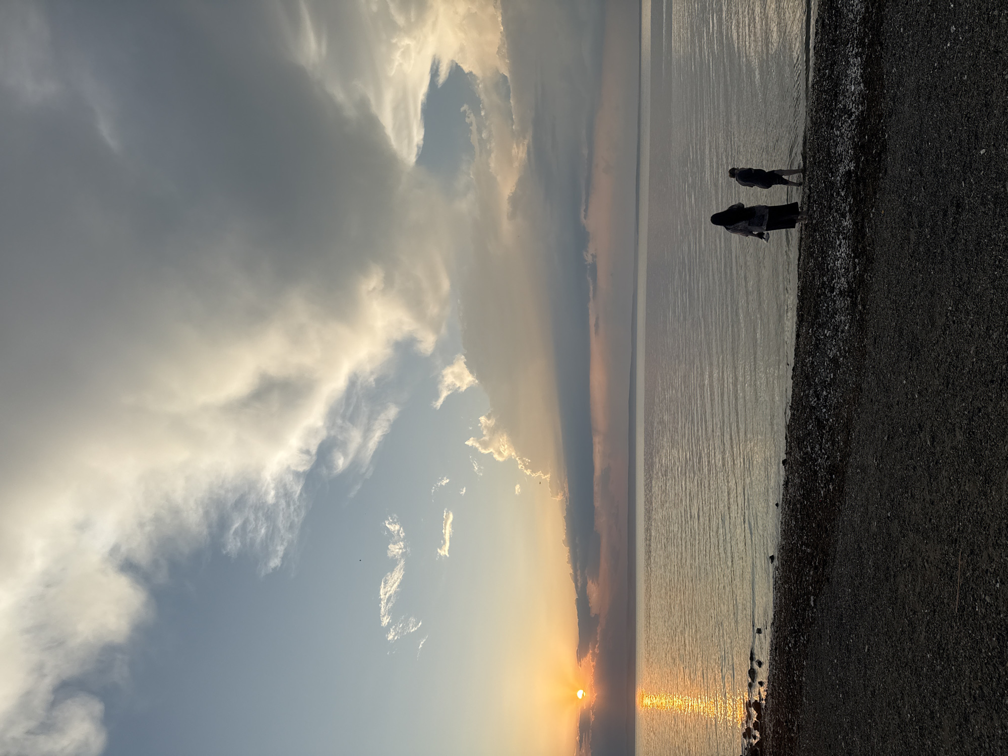 The image captures a tranquil scene at a beach in Vladivostok, Russia, likely during a sunset. The sky is dramatic with a mix of dark, heavy clouds and bright patches where the sun's rays break through, suggesting a dynamic weather event. The sun itself is low on the horizon, casting a warm, golden glow that reflects on the gently rippling water.
The foreground is a dark, pebbly beach, with the water's edge marked by small stones. Two figures are visible near the water, silhouetted against the bright expanse of the sea. One person appears to be wearing a dark top and lighter bottoms, while the other is in a dark outfit. They are standing still, seemingly observing the sunset or enjoying the peaceful atmosphere.
The overall mood is serene and contemplative, with the interplay of light and shadow creating a sense of depth and vastness. The calm water and the solitary figures suggest a moment of quiet reflection. The weather appears to be transitioning, with the sun's emergence from behind the clouds adding a touch of hope and beauty to the otherwise moody sky. There is no visible text in the image.
