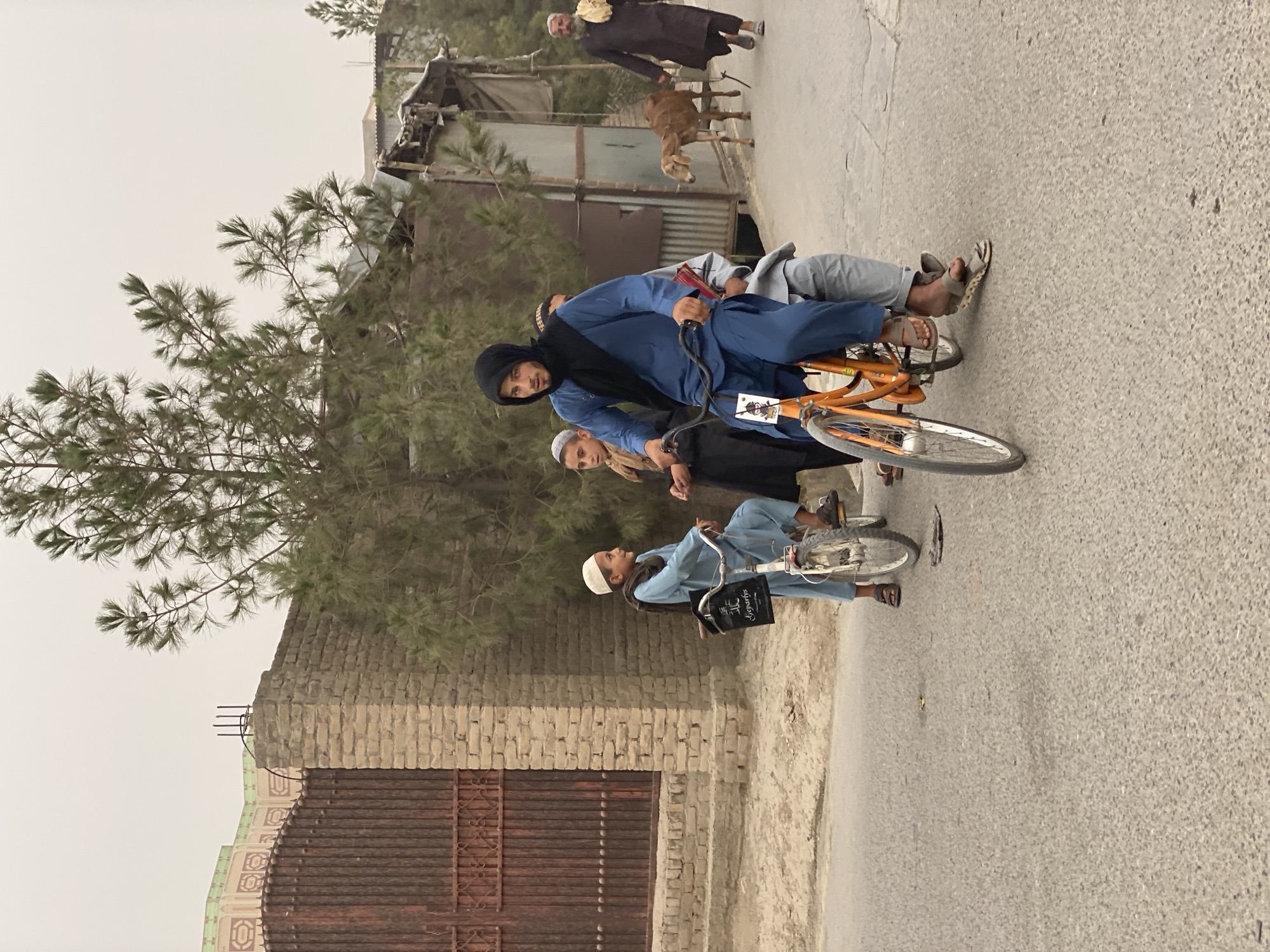 In a street scene in Kandahar, Afghanistan, a group of individuals are captured. The foreground is dominated by a young man wearing a blue shalwar kameez and a black headscarf, riding an orange bicycle. He is looking towards the camera. A child in a light blue shalwar kameez and a white taqiyah is seated behind him on the bicycle, and another child in a black shalwar kameez and white taqiyah is standing next to the bicycle. Further to the right, a man in a dark brown shalwar kameez walks with a stick, accompanied by a brown goat on a lead. In the background, a brick wall with an ornate, rusty metal gate stands to the left, partially obscured by trees. Buildings with corrugated metal roofs and weathered walls are visible further back, suggesting an urban or semi-urban environment. The ground is paved with asphalt. The overall atmosphere is overcast.