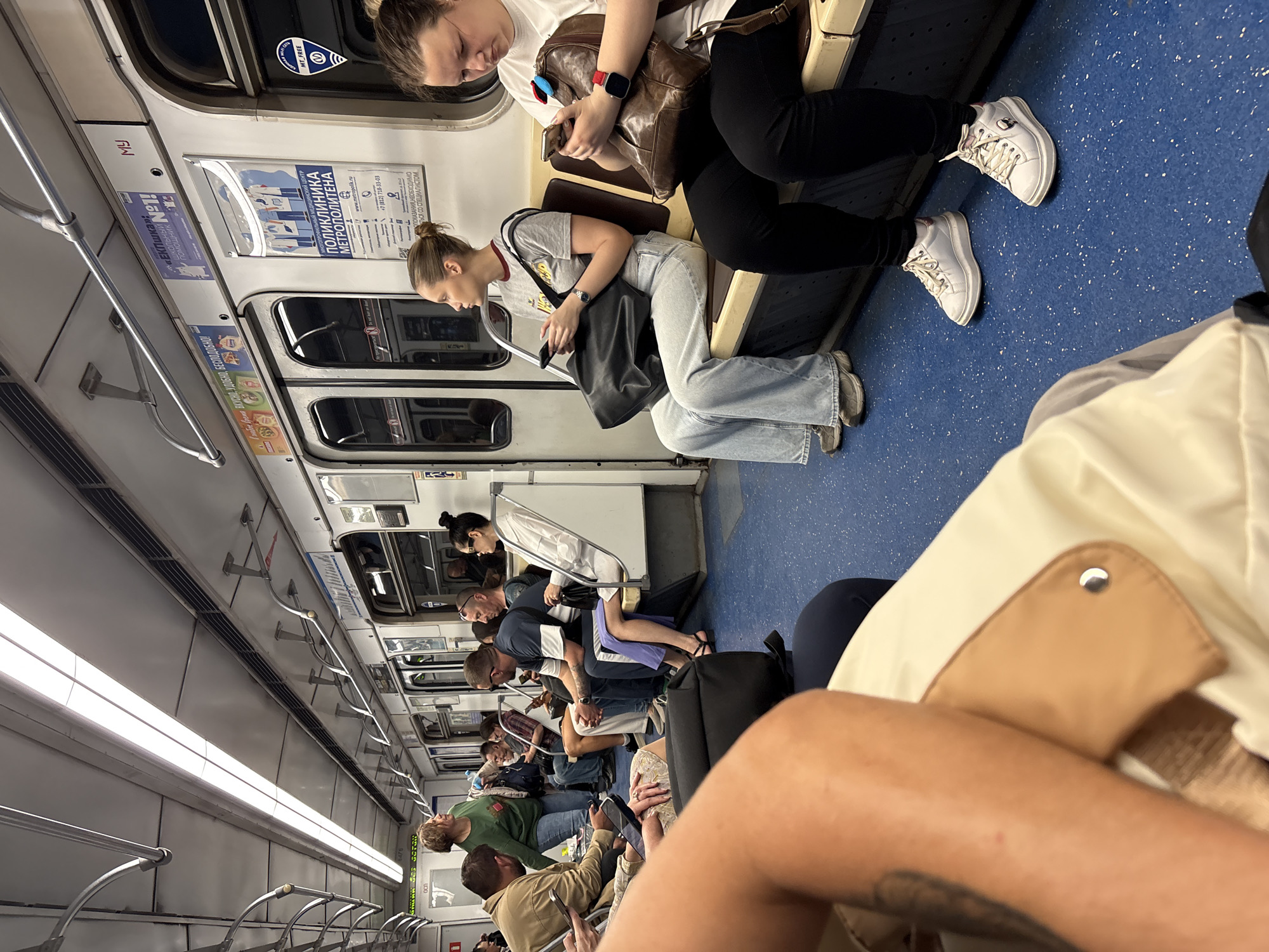 The image depicts the interior of a subway car, likely in an urban setting. Occupants are seated on benches along the sides, many preoccupied with mobile devices. Advertisements are visible on the walls of the car, along with safety signage. Seated passengers are of various ages.