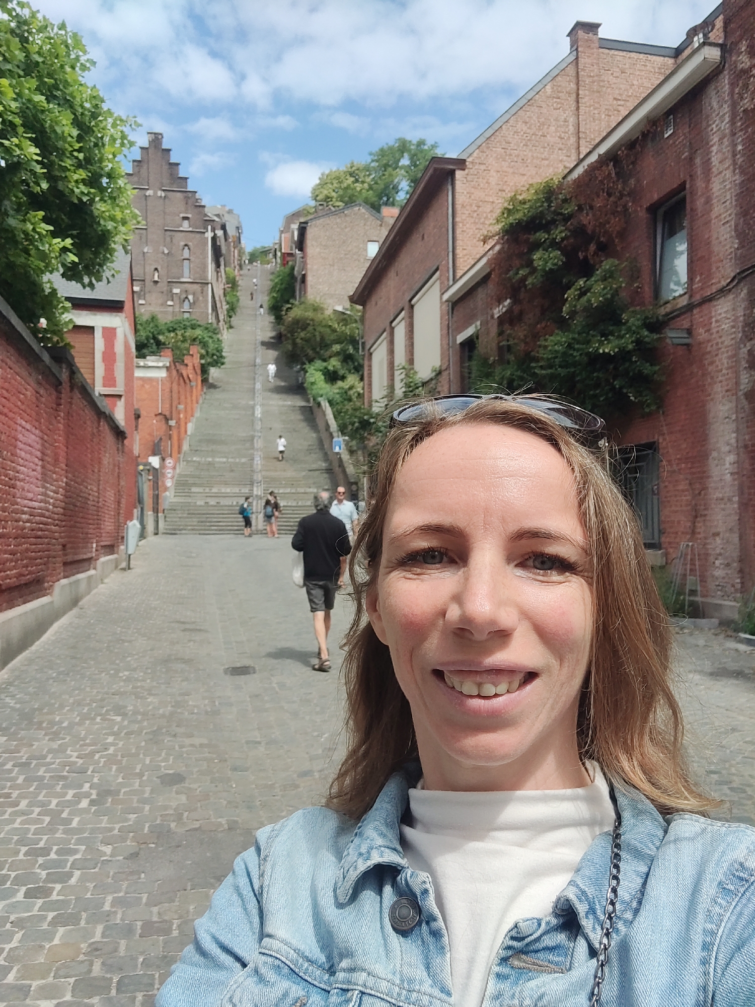 A selfie is taken by a Caucasian woman with shoulder-length blonde hair, wearing a light blue denim jacket over a white turtleneck. She is smiling and looking at the camera. Behind her, a cobblestone street slopes upwards, lined with red brick buildings and a tall, stone staircase. Several people are visible on the stairs and street. The woman is positioned in the foreground, with the city street and architecture filling the background. The location is the Montagne de Bueren staircase in Liège, Belgium.