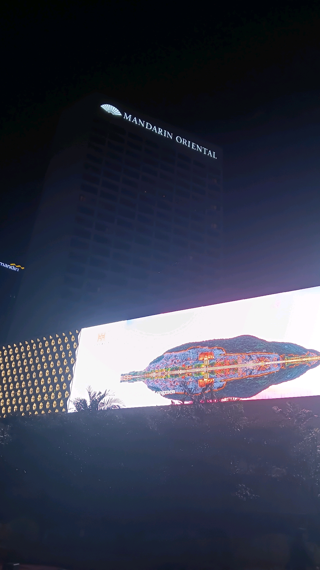 This is a nighttime, medium-angle shot of the Mandarin Oriental hotel in Central Jakarta, Indonesia. The hotel's iconic fan logo is illuminated in white above the hotel's name, also in white, "MANDARIN ORIENTAL". A large LED screen is positioned below the hotel, displaying a vibrant image of a city built into a mountainside, reflected in a body of water. Palm trees and other foliage are visible in front of the screen. The scene is dark, with artificial lights illuminating the hotel and the LED display.
