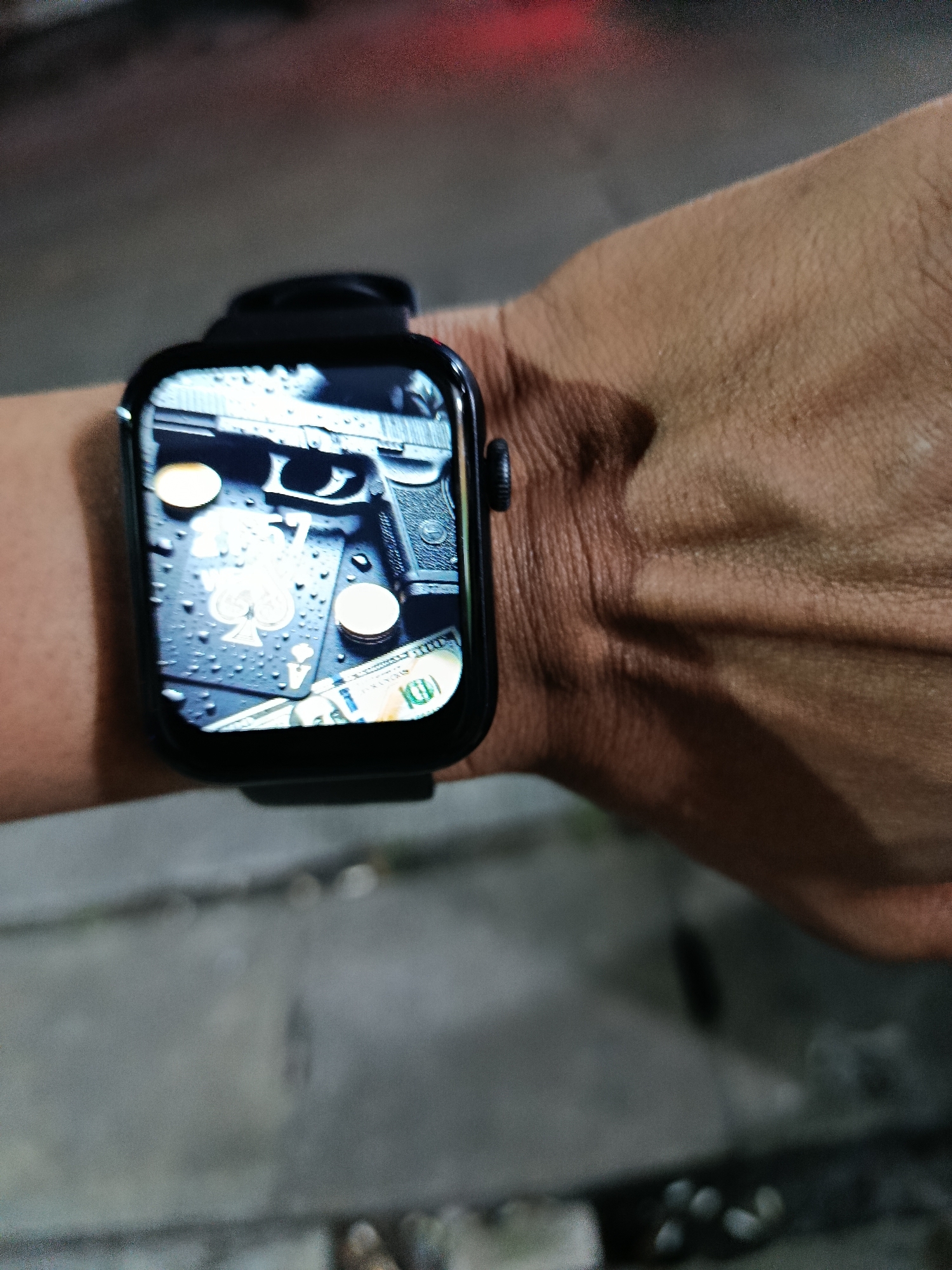 A close-up shot features a person's wrist wearing a smartwatch. The smartwatch displays a wallpaper with a handgun, playing cards, coins, and US dollar bills. The background is blurred, showing a concrete-like surface and a hint of red light, possibly from a vehicle or ambient lighting. The image appears to be taken in an outdoor urban environment. The specific location in Depok, Indonesia, cannot be determined from the visual information alone.