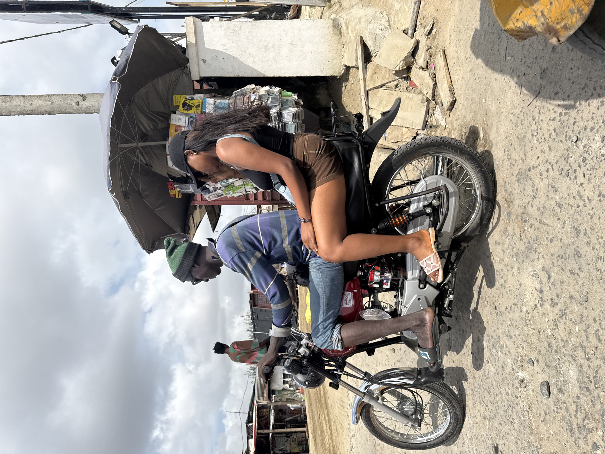An outdoor, medium shot captures two individuals riding a red motorcycle on a dirt road in Ado, Nigeria. The rider, a male wearing a green beanie and a blue and purple striped shirt, is in the foreground, facing left. His legs are bare, and he wears dark sandals. The passenger, a female, sits behind him, facing left, wearing a black tank top, brown shorts, and a black baseball cap. Her left arm rests on the rider's shoulder, and she holds a black handbag on her left side.
Behind the motorcycle, a dark grey umbrella covers a stall stocked with various items. A white pillar stands to the right of the stall, and behind that, a larger white structure is visible. The background shows more stalls with corrugated metal roofs and a cloudy sky.
In the middle ground, a person wearing a colorful shirt stands facing away from the camera. The road surface is a mixture of dirt and small rocks. The scene appears to be a bustling market area or a roadside commercial zone.