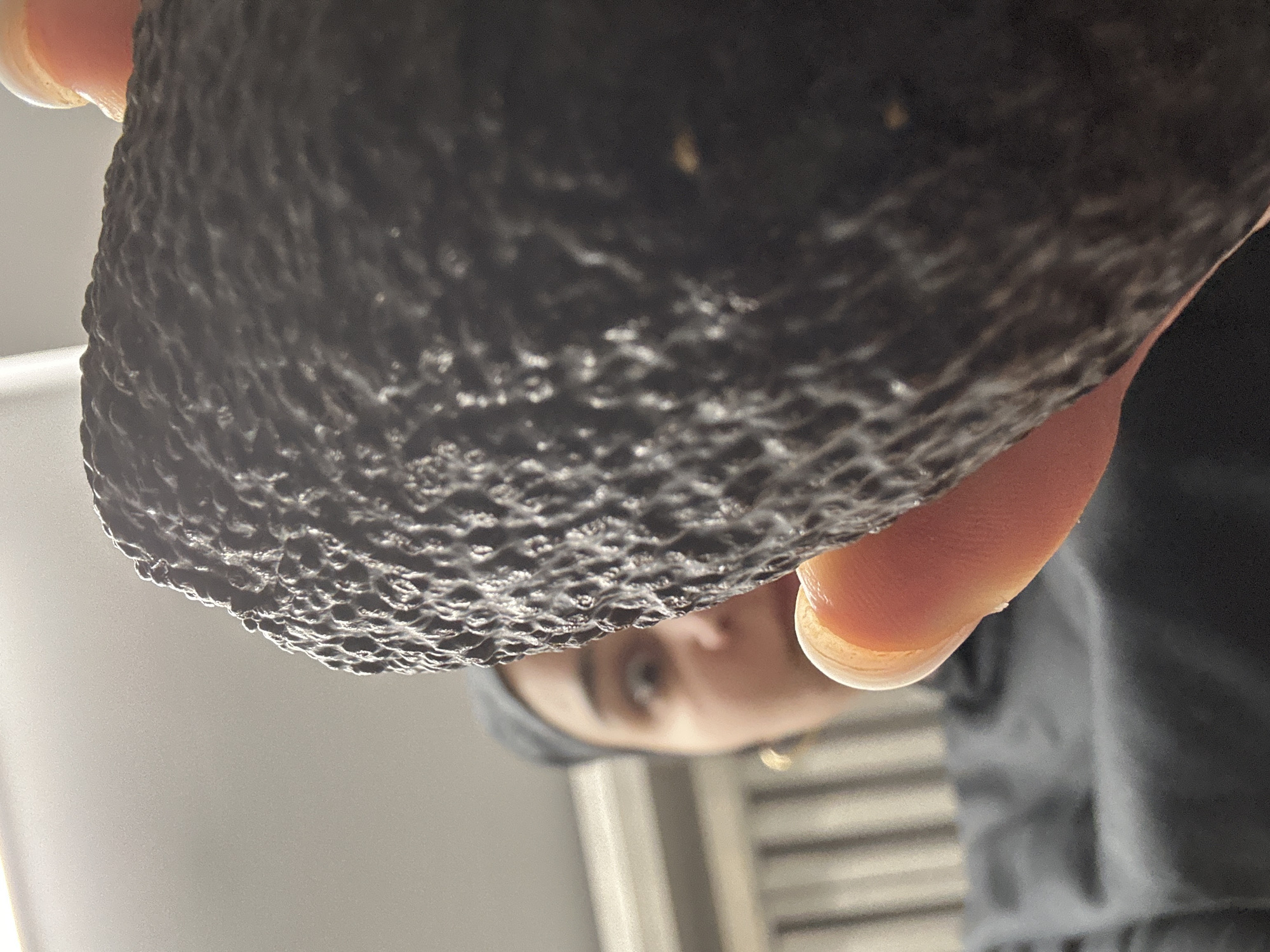 A close-up, upward-angled view shows a dark, rough-textured avocado being held by a person. The person's hand is visible, with a portion of a finger and fingernail in the upper right. Behind the avocado and hand, the person's face is partially visible, including an eye and part of the nose. They are wearing a dark hooded garment. The background consists of a white or light gray wall with a set of horizontal lines or blinds. There is no information available to determine if this photo was taken in Burgas, Bulgaria.