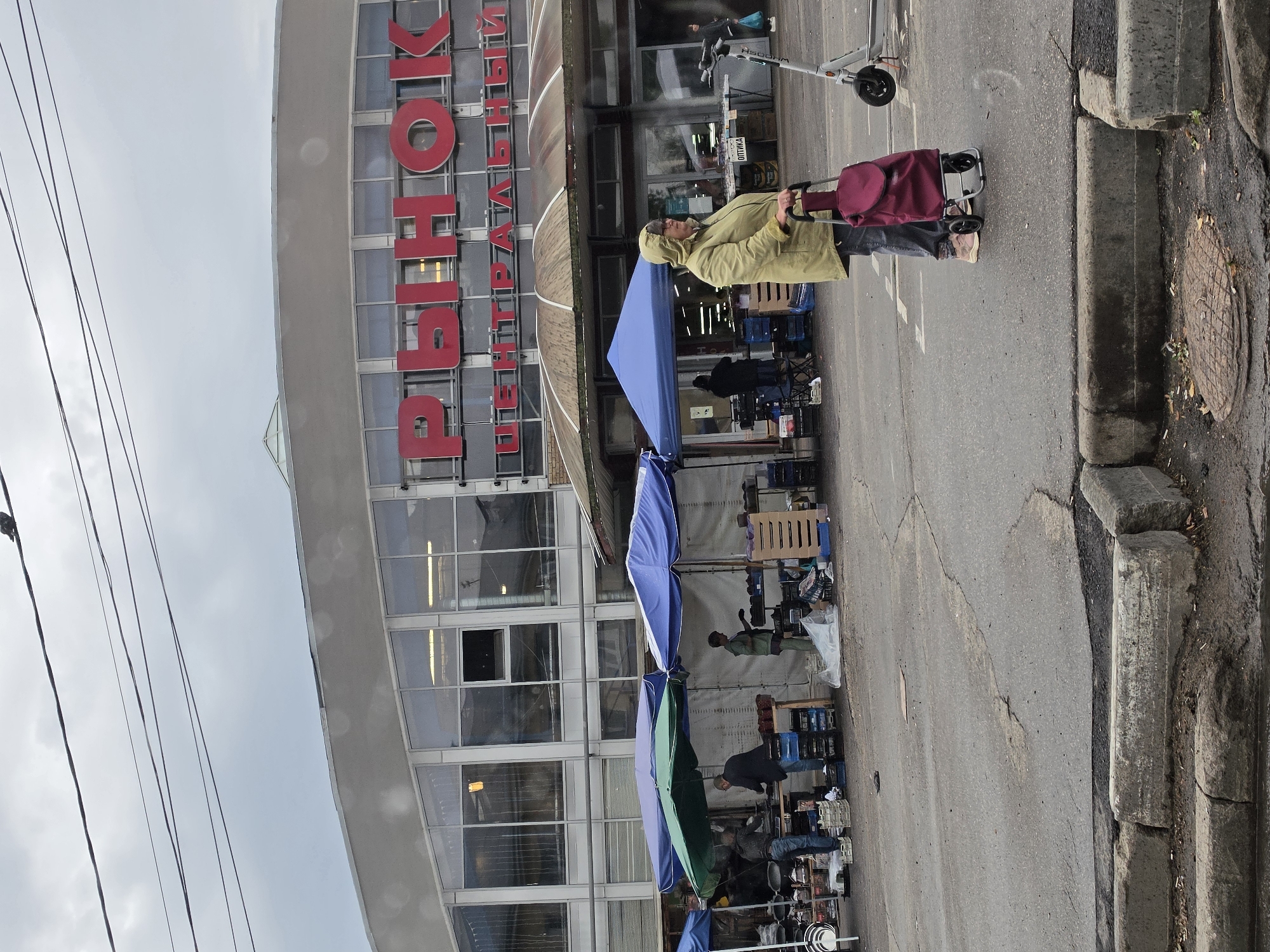 A woman in a tan hooded jacket and jeans is standing on a cracked asphalt surface, holding a red rolling shopping cart with a dark red bag. She is looking to her right. Behind her, to her left, are market stalls with blue and green canopies, displaying various goods. Several people are visible in the background, tending to the stalls. The building behind the stalls has large windows and features prominent red Cyrillic lettering that reads "РЫНОК" (Rynok, meaning "Market") and "ЦЕНТРАЛЬНЫЙ" (Centralny, meaning "Central"). A white electric scooter is parked to the right of the woman. The sky is overcast. The image appears to be taken in Nizhny Novgorod, Russia, specifically at the Central Market.