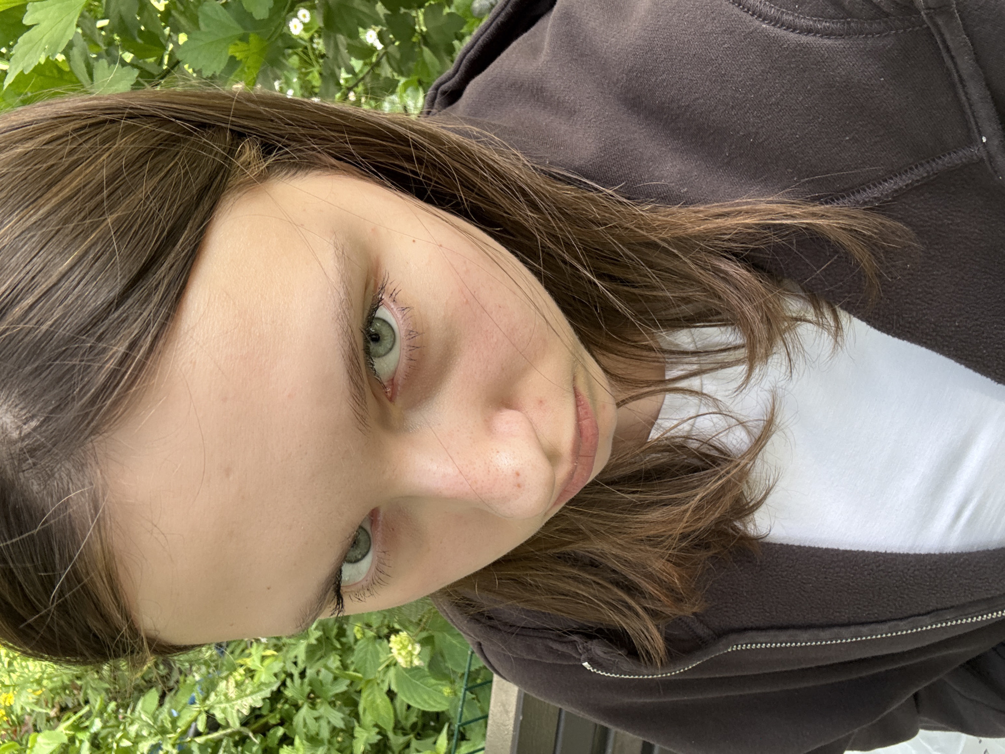 A young person with shoulder-length, brown hair and green eyes is captured in a close-up, slightly tilted shot. They are wearing a dark brown hooded zip-up jacket over a white t-shirt. The background consists of lush green foliage and some small white flowers, suggesting an outdoor setting with ample vegetation. The lighting appears to be natural, indicating daytime. The person's expression is neutral, with their gaze directed slightly away from the camera. The image is set outdoors, with greenery and hints of what might be a fence or railing in the lower portion of the frame. There is no visible text in the image.