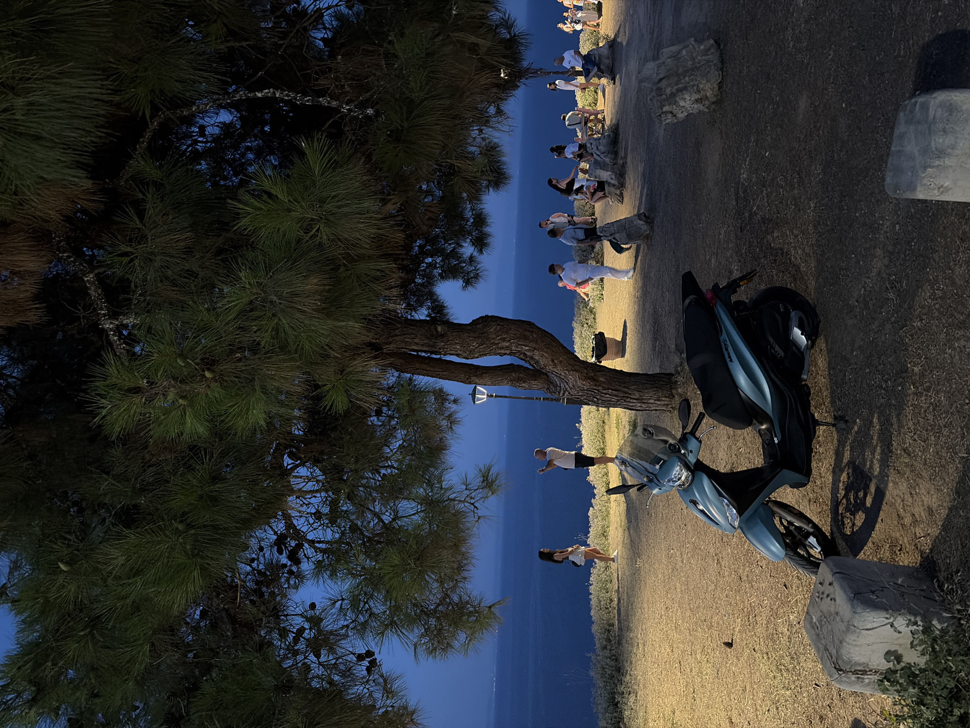 The image depicts a dusk scene in Kassandra Municipal Unit, Greece, with a view of the Aegean Sea. In the foreground, a teal-colored scooter is parked on a patch of dry grass and dirt, with a large, rough-hewn stone block to its left. Beyond the scooter, a group of people are gathered on a slightly elevated area overlooking the sea. Several individuals are standing or sitting on rocks and a wooden bench, some appearing to be engaged in conversation or enjoying the view. A lamppost with a warm light is visible near a tree trunk on the right side of the frame. The sea stretches out to the horizon, with distant lights indicating a town or settlement. The sky is a deep twilight blue, with the branches of pine trees, laden with cones, forming a canopy over the scene. The overall atmosphere is one of a relaxed evening gathering in a coastal location.
